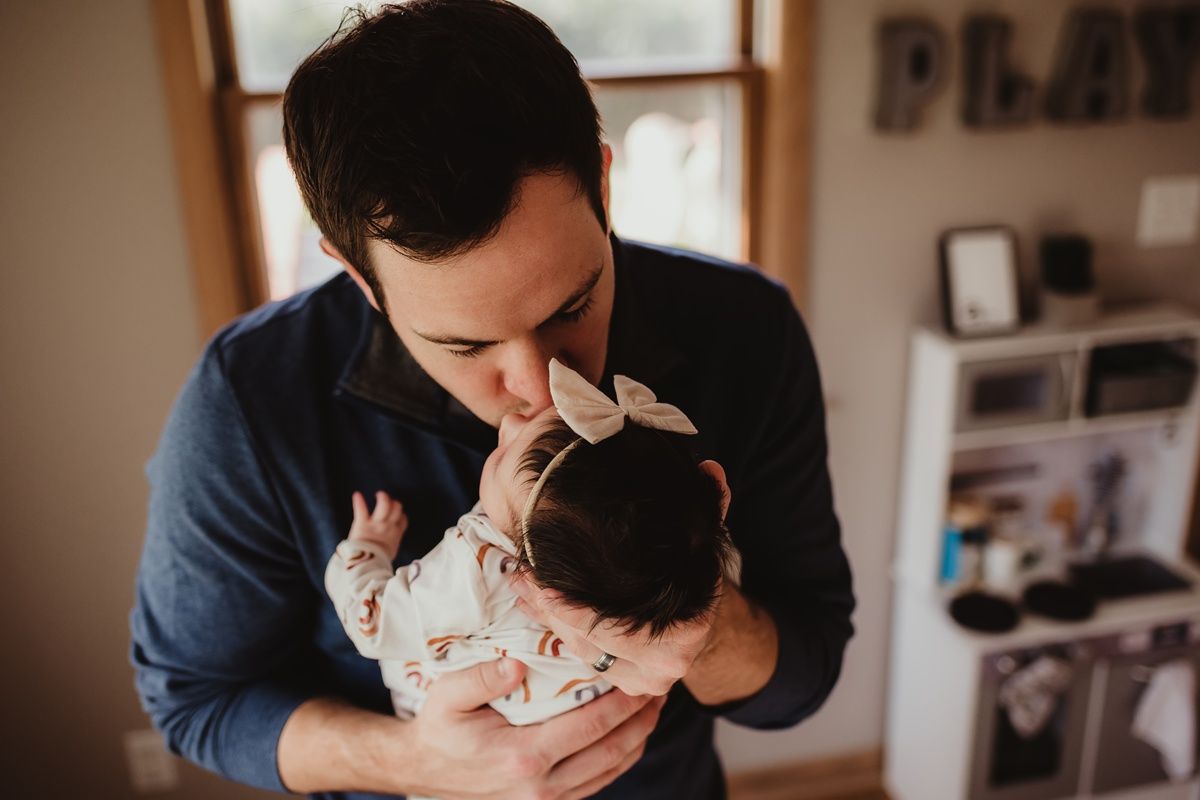 Parent in blue sweater lovingly kisses newborn's head near window.