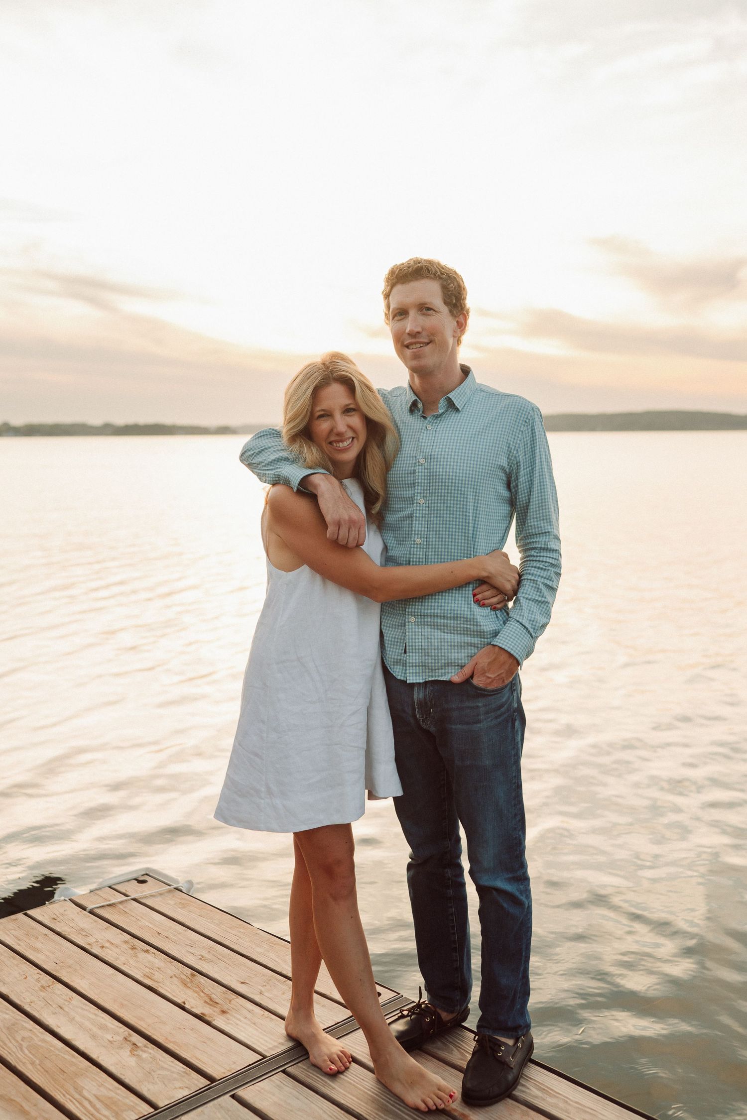 A couple embraces on a wooden dock during a golden sunset over the water.