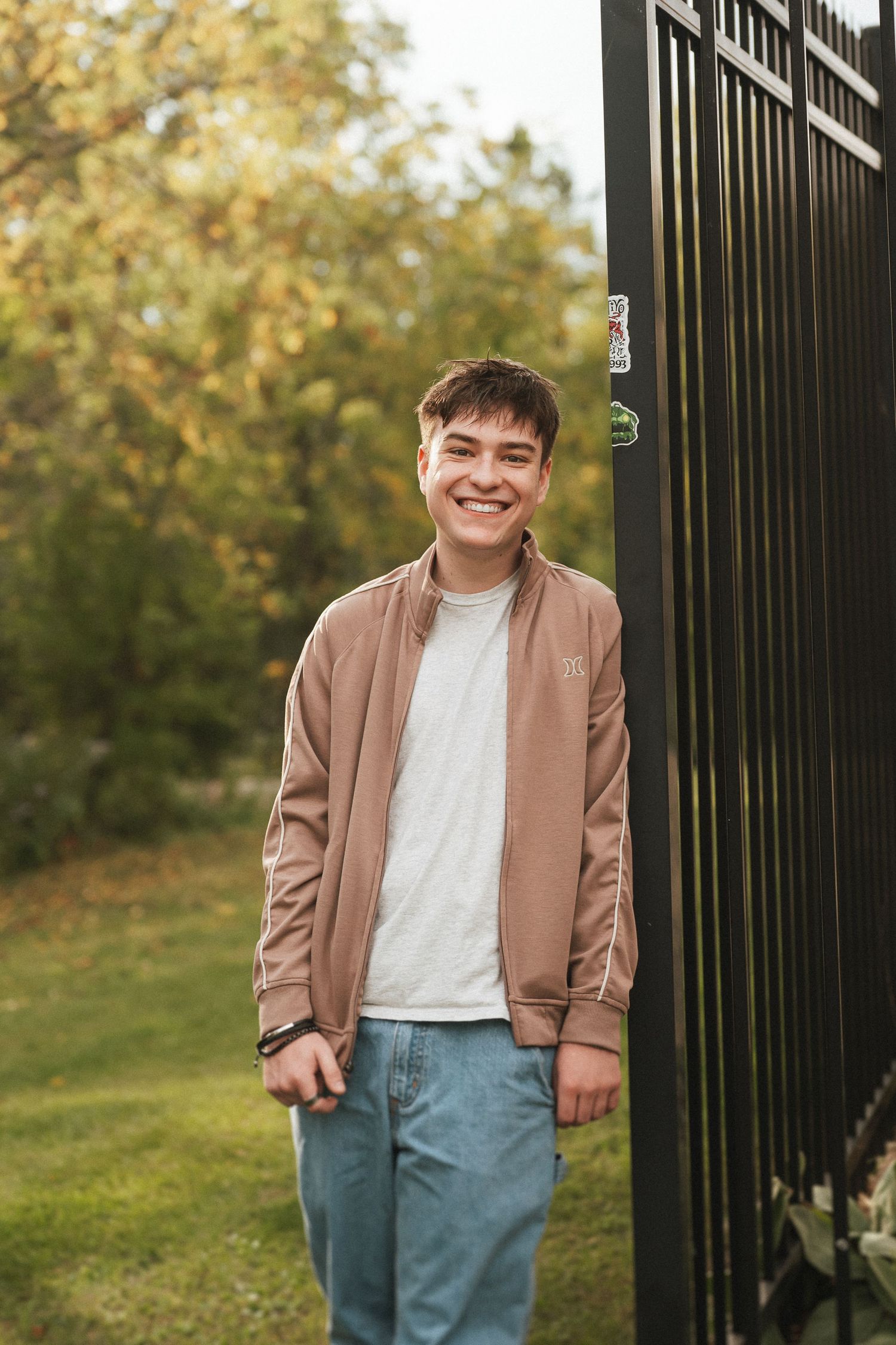 A person wearing a tan blazer and light colored top poses against a black metal fence with autumn foliage in background.