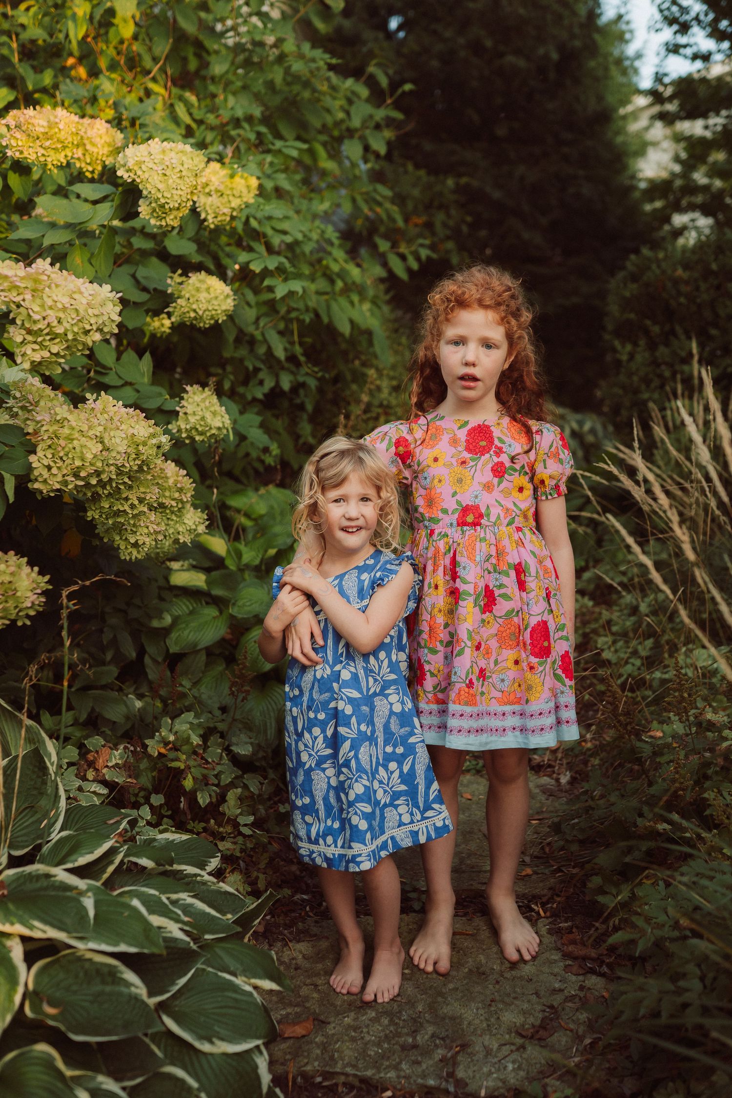 Children in floral dresses standing among garden flowers and hostas.