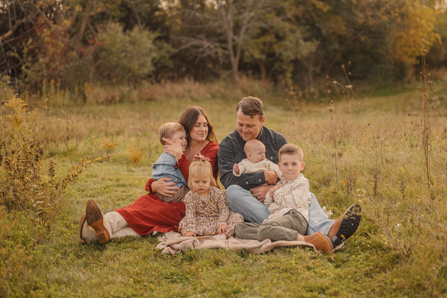 A group sits together on a blanket in a golden autumn field during sunset for a casual outdoor portrait session.