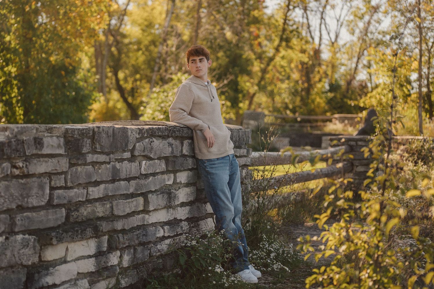 A person sits on a stone wall during golden hour autumn photography session in a park setting.