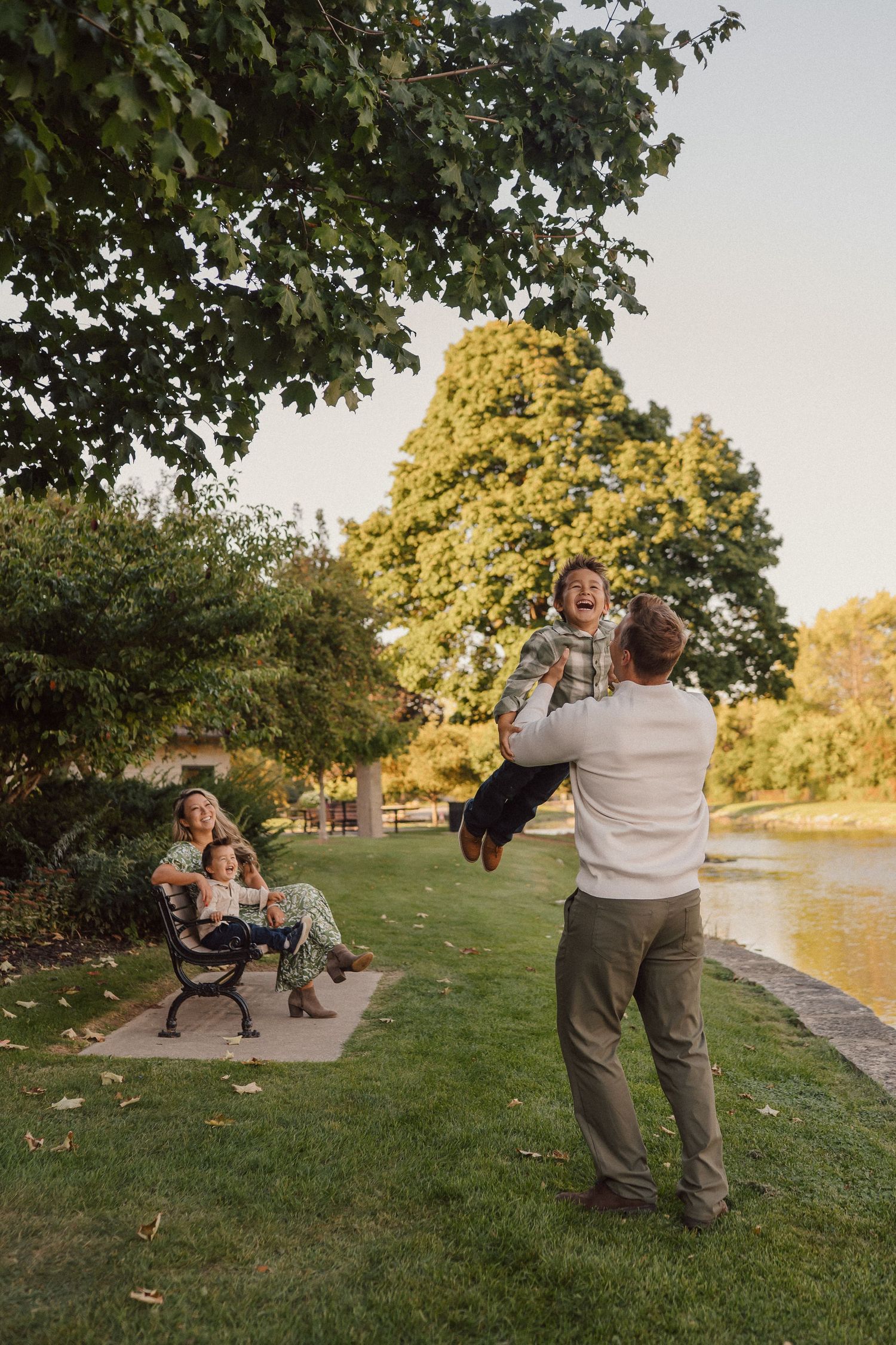 Family enjoying a sunset moment by a pond in a park with trees and a bench.