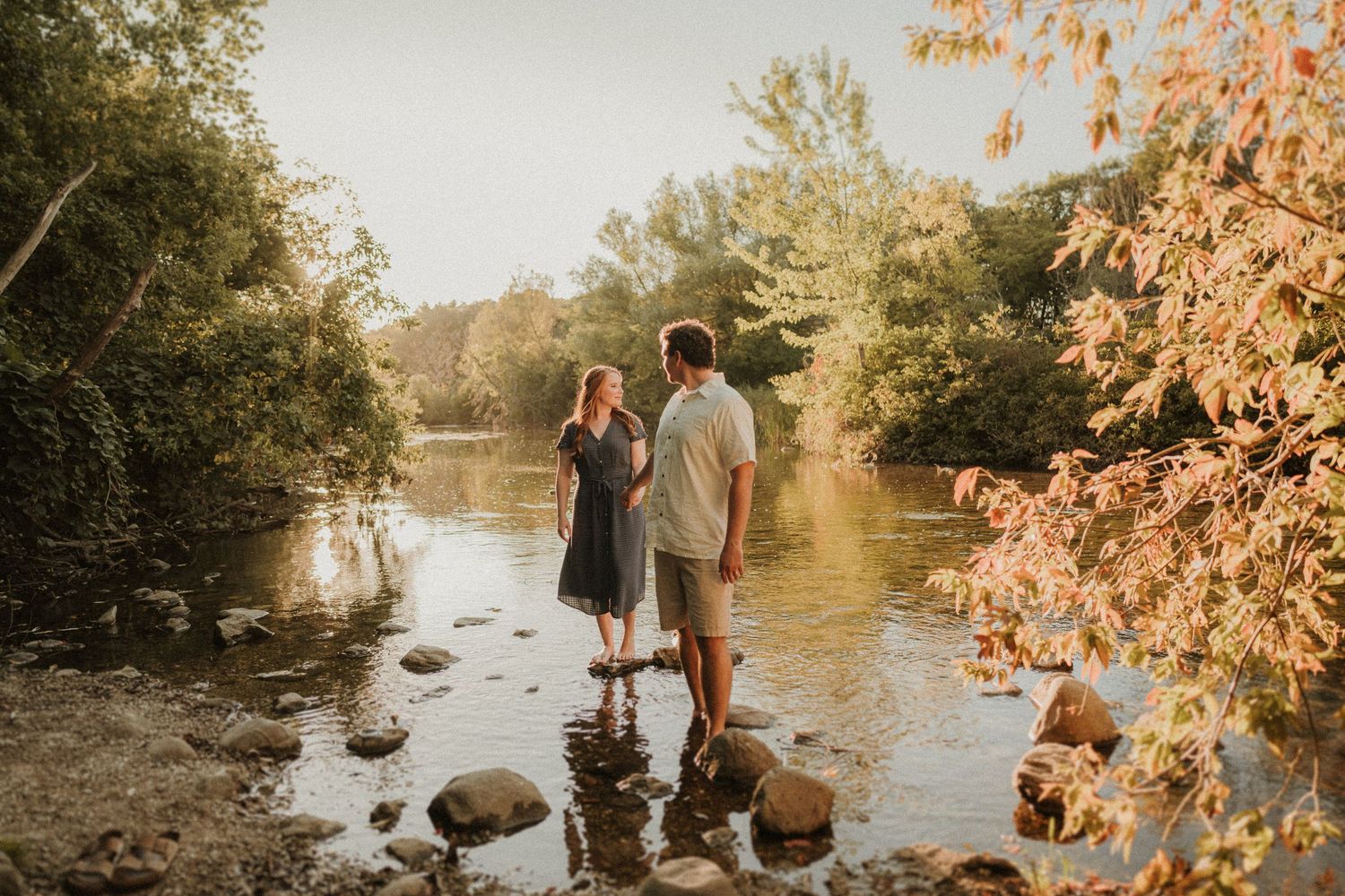 Couple standing together on rocks by a river during golden hour with autumn trees in background.