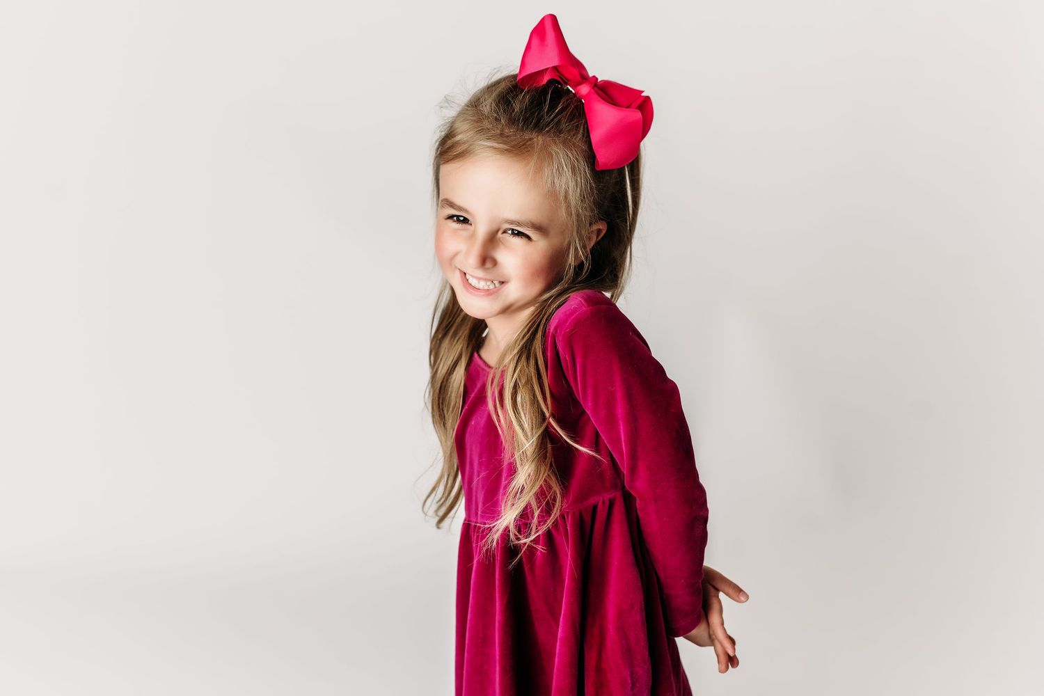 Child in burgundy dress and pink bow smiling cheerfully against white background.