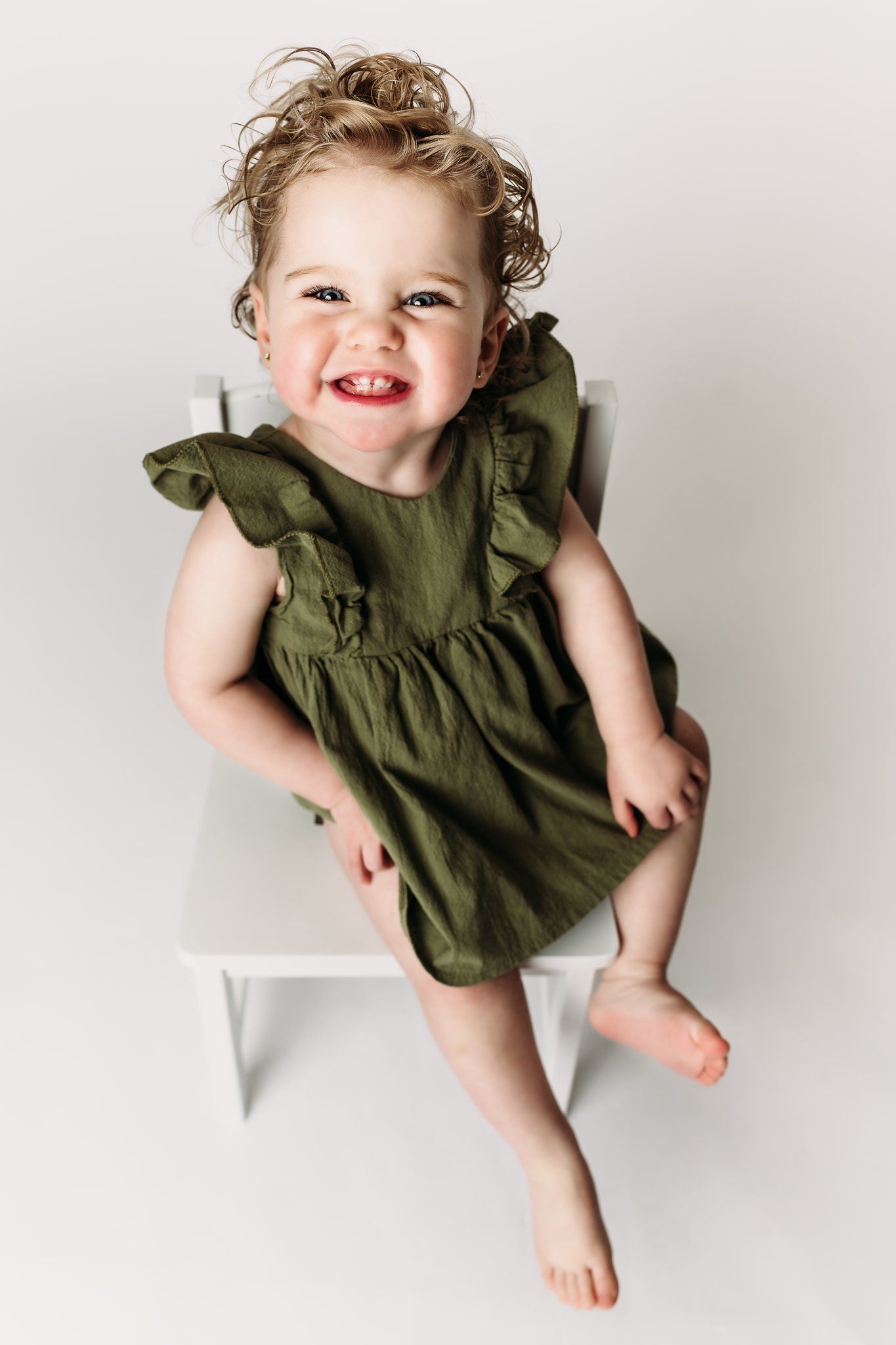 Joyful child in green dress sits on white chair with dangling feet.