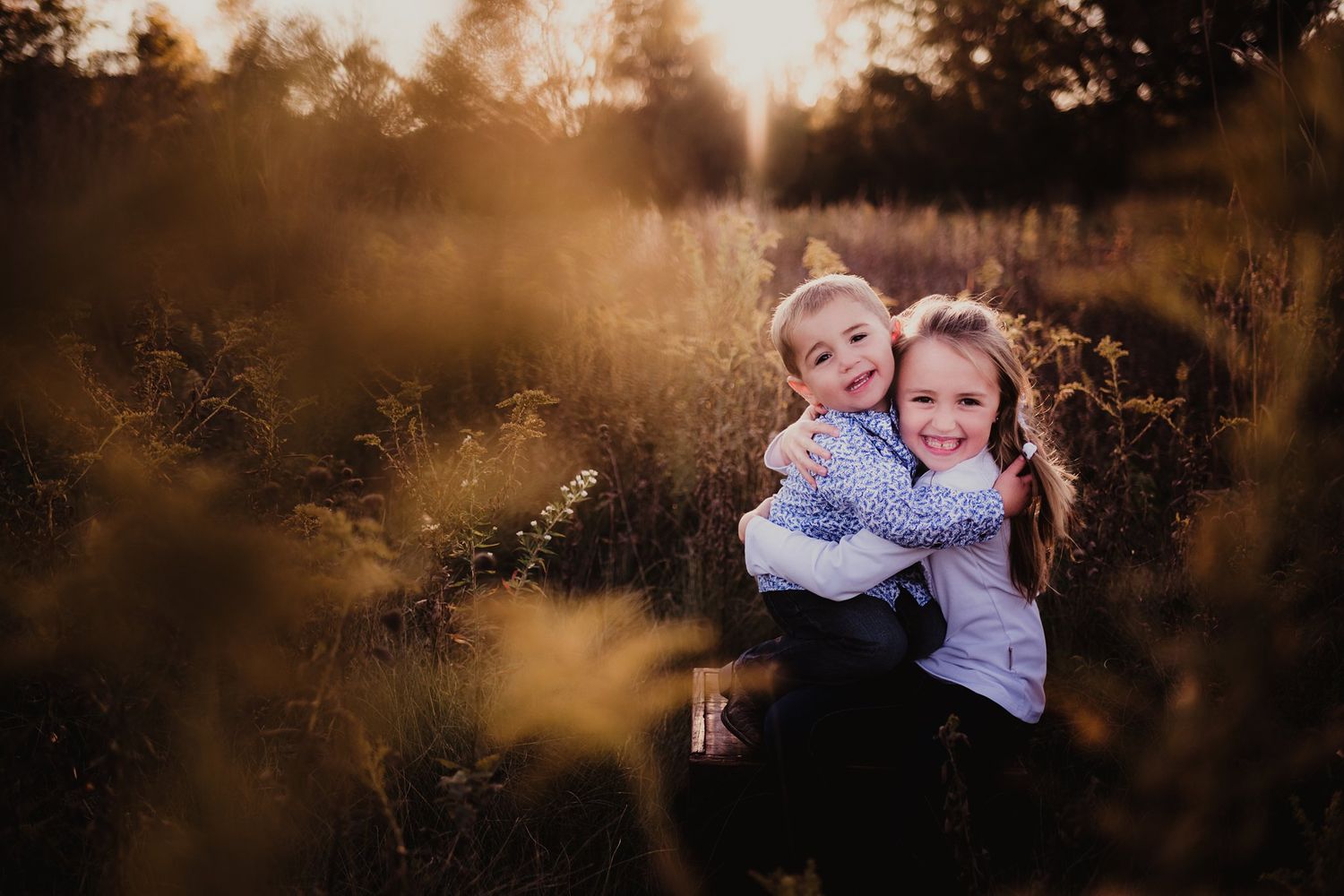 Children hugging and smiling in tall grass during golden hour.
