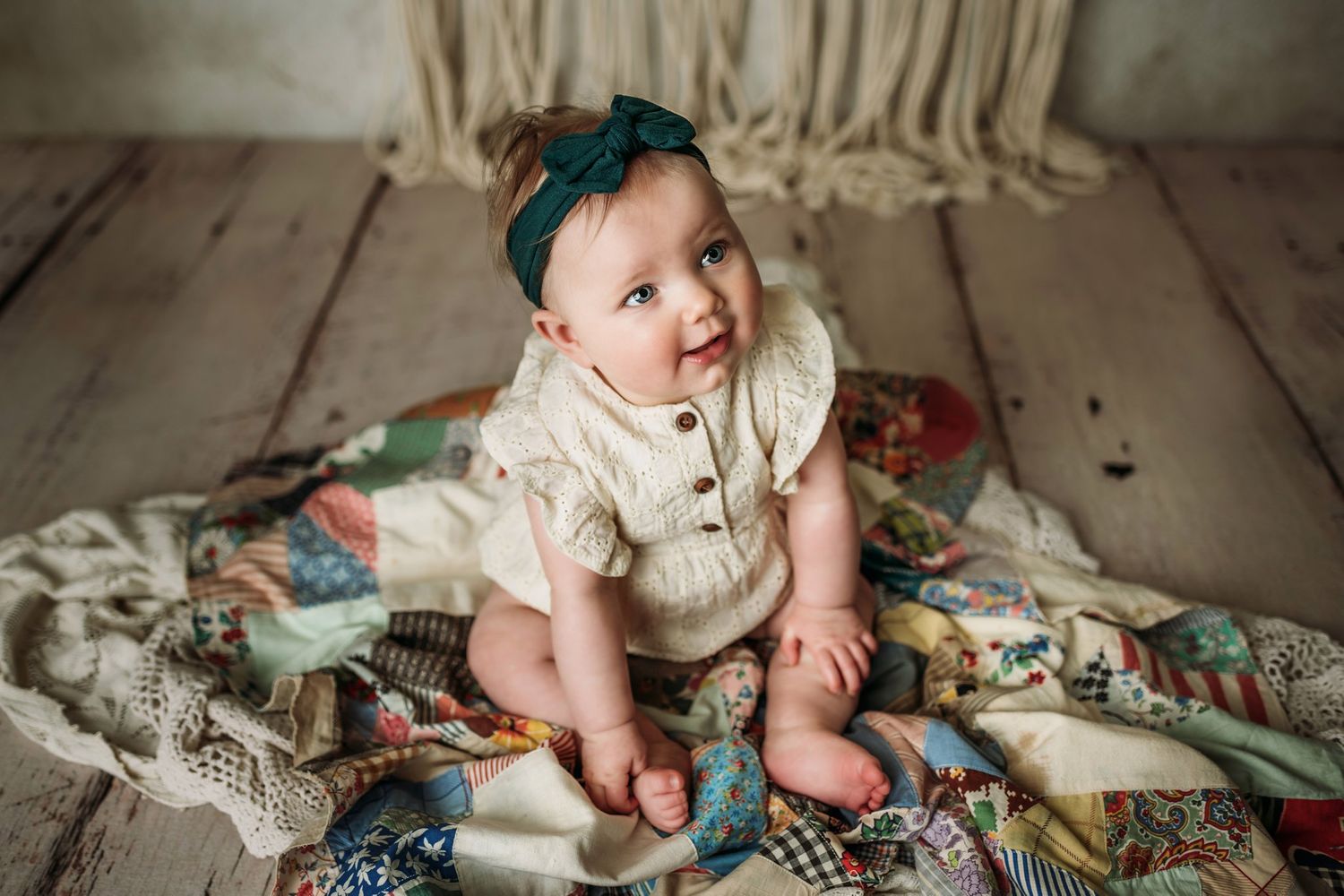 Smiling baby in vintage-style cream outfit sits on patterned quilt and lace.