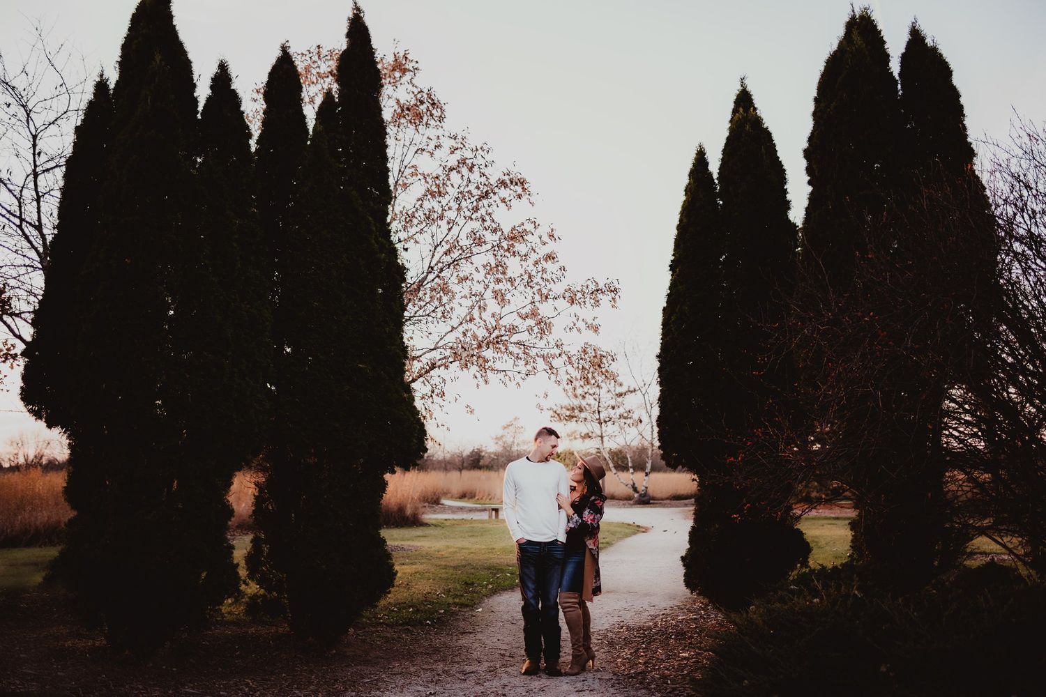 Silhouetted couple standing between rows of tall cypress trees at dusk.