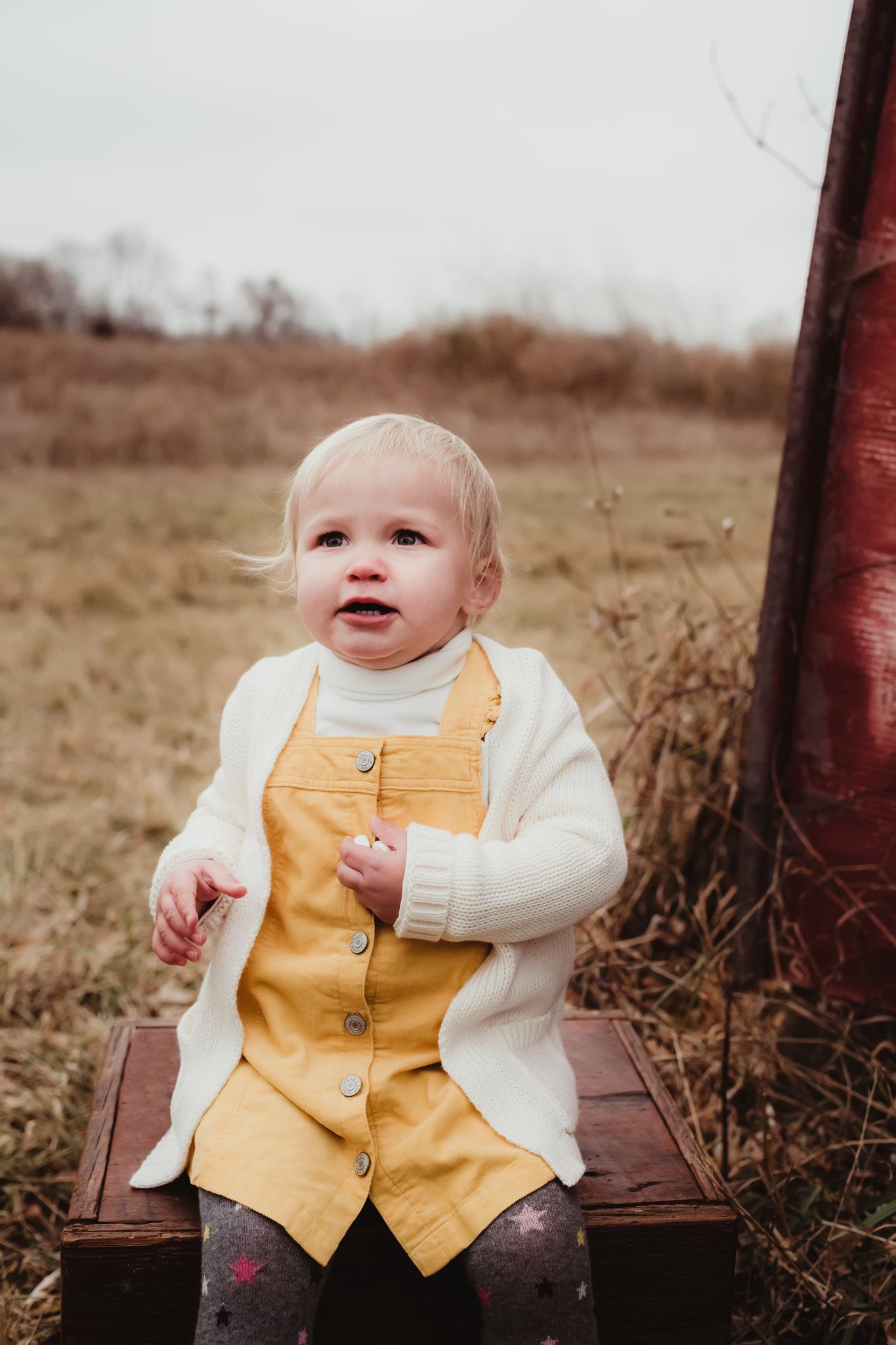 A toddler in yellow overalls sits on wooden steps next to a red wall outdoors.