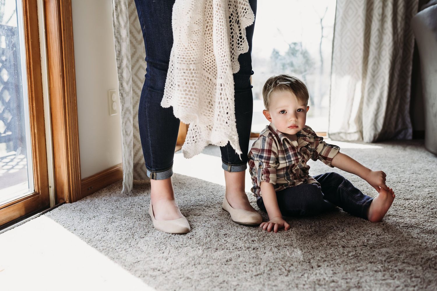 Young child in plaid shirt sits on carpet near window with lace curtains.