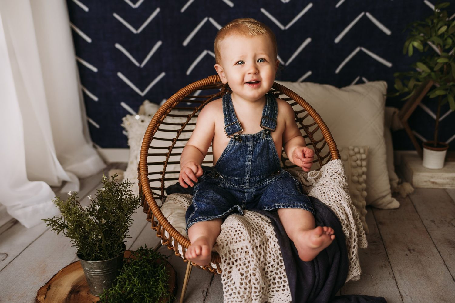 Laughing baby in denim overalls poses in rattan chair against navy patterned wall.