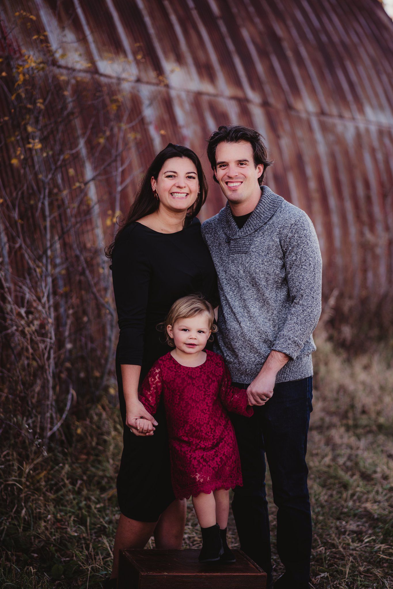 A family portrait taken against a rustic red barn wall with coordinated fall outfits.