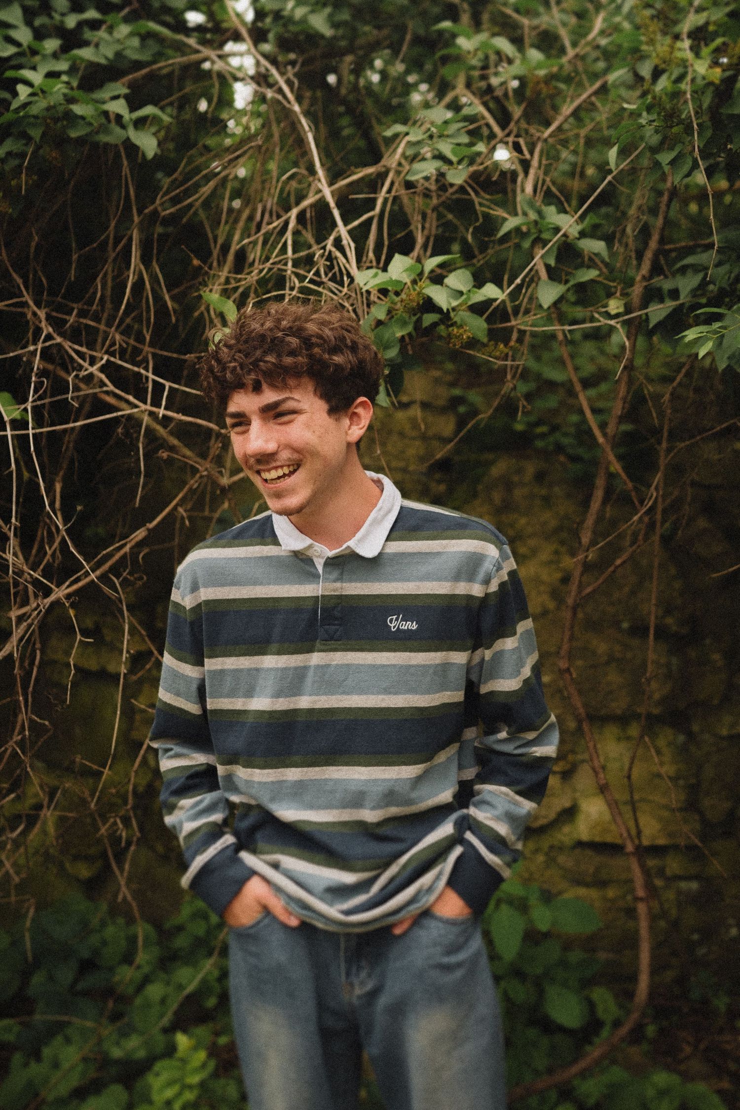 Casual outdoor portrait of someone wearing a striped sweater and jeans standing against leafy foliage.