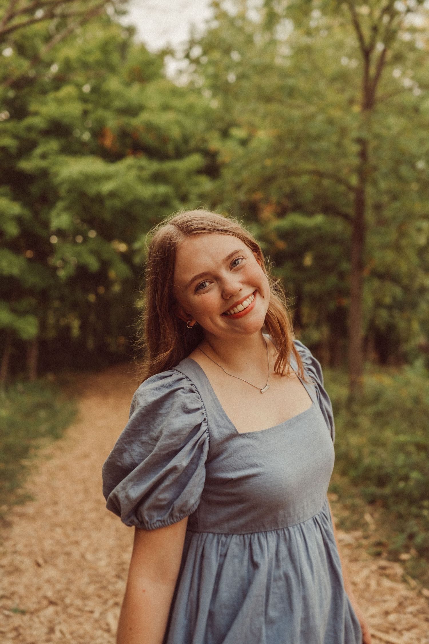 A joyful portrait in a blue dress with puff sleeves standing on a wooded forest path.