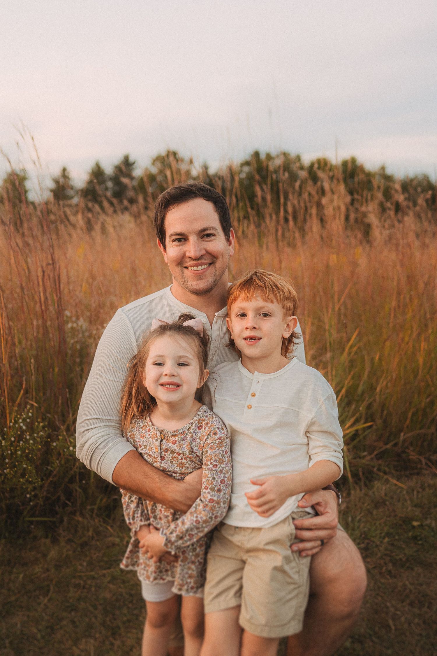 Family portrait in a golden field with tall grass and trees at sunset.