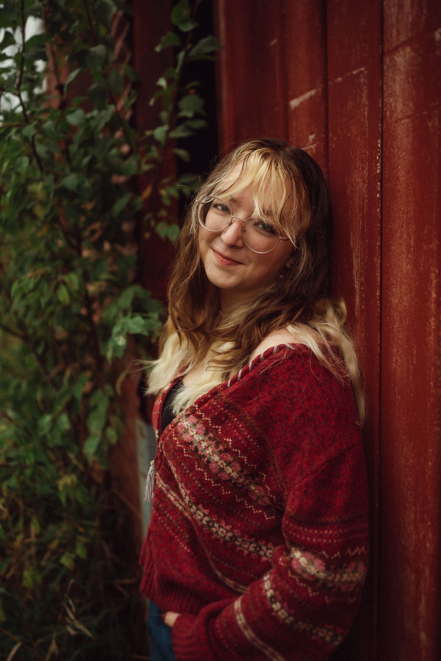 A cozy portrait against a red barn wall featuring glasses and a festive red knit sweater with fair isle patterns.