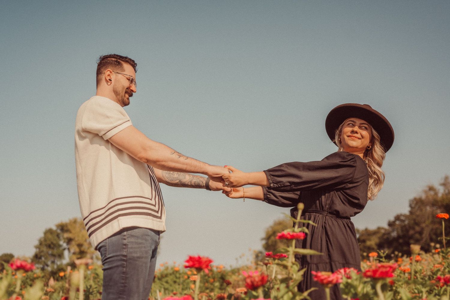 A couple holds hands while standing in a vibrant field of red flowers during sunset.