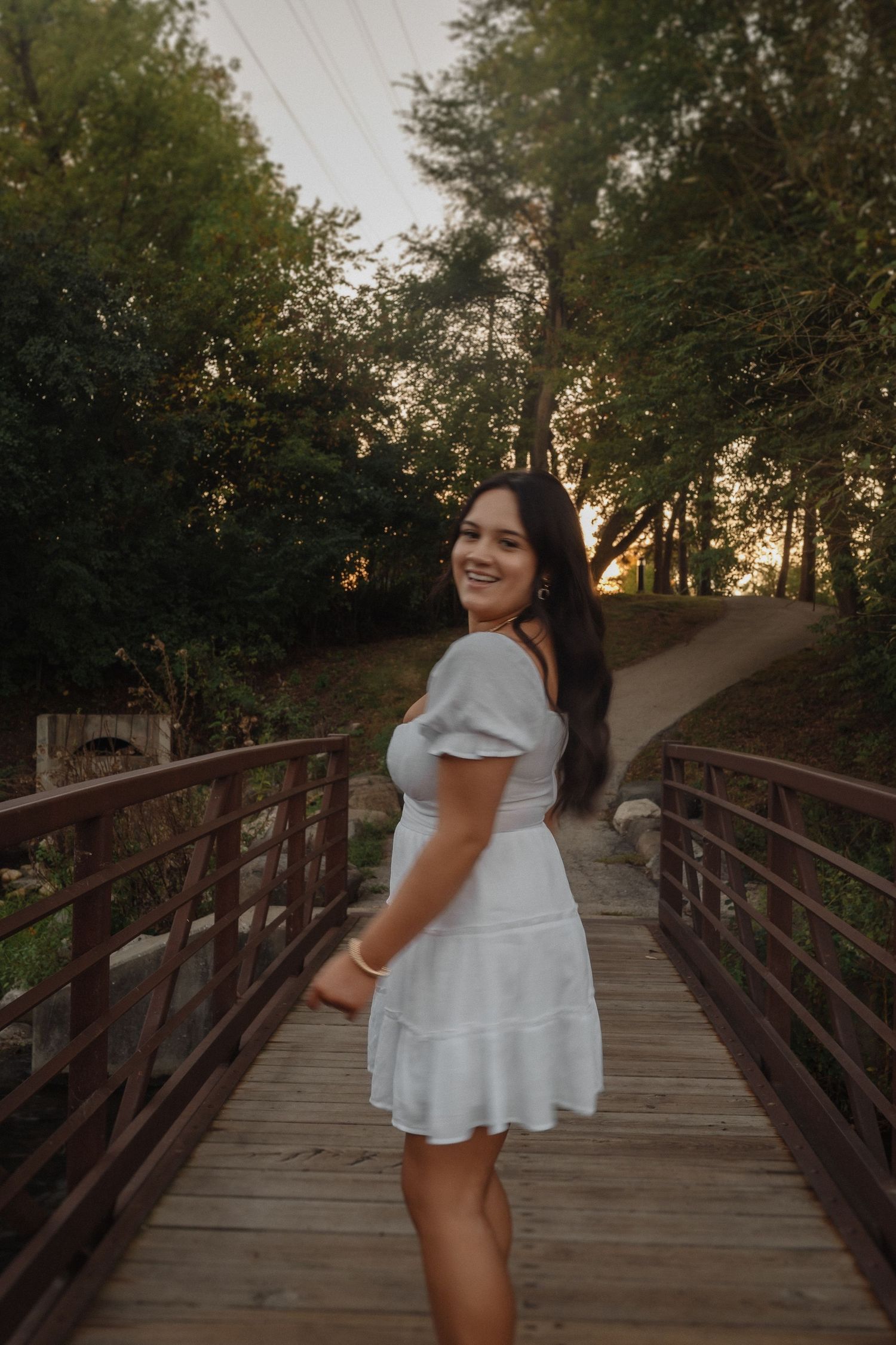 A person in a white summer dress poses on a wooden bridge surrounded by trees during golden hour.