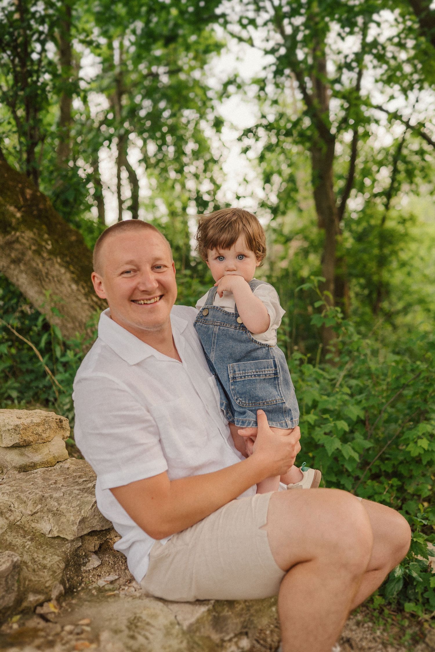 A heartwarming outdoor portrait of an adult and small child sitting together on a rocky outcrop in a wooded setting.