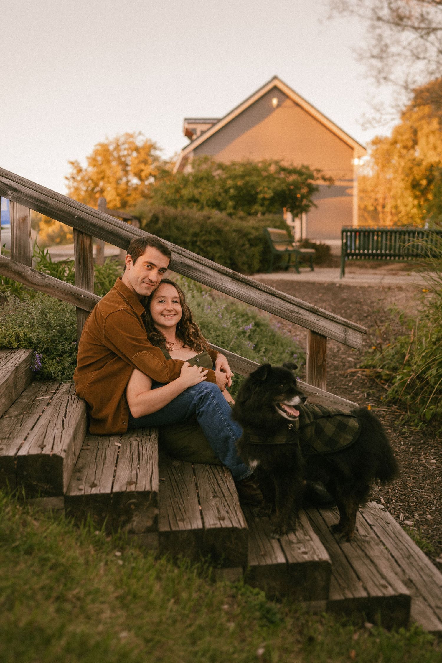 A couple sits on wooden stairs outdoors during autumn with their black dog beside them.