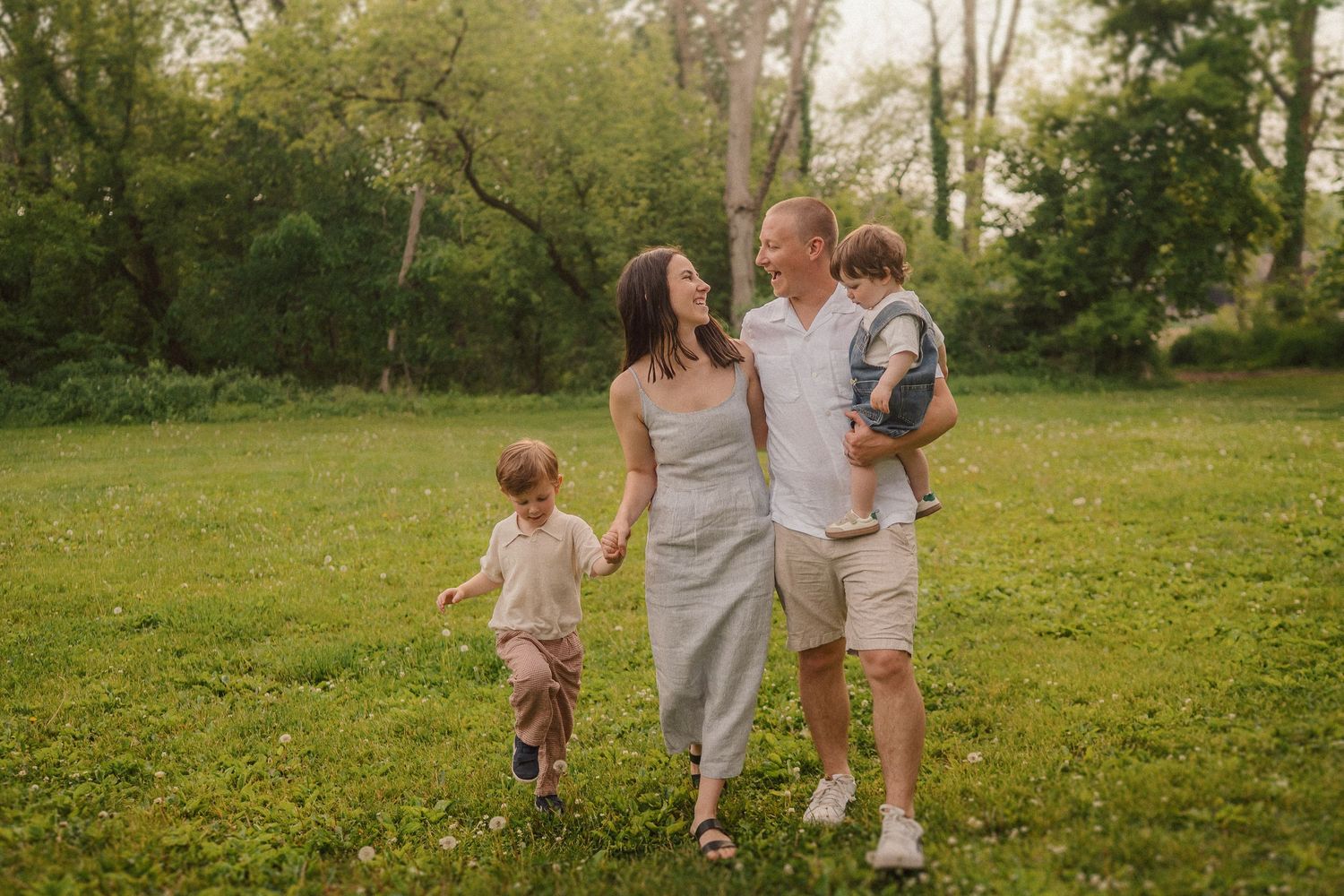 A happy family walks together through a lush green park filled with dandelions and spring flowers on a sunny day.