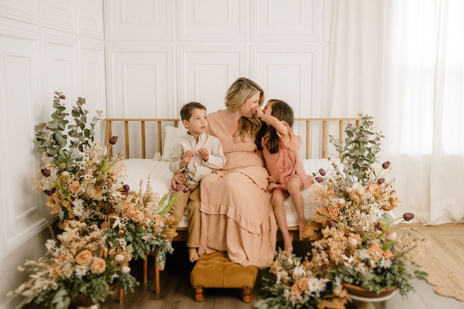 Three people sit together on a wooden bench surrounded by floral arrangements in a bright studio setting.
