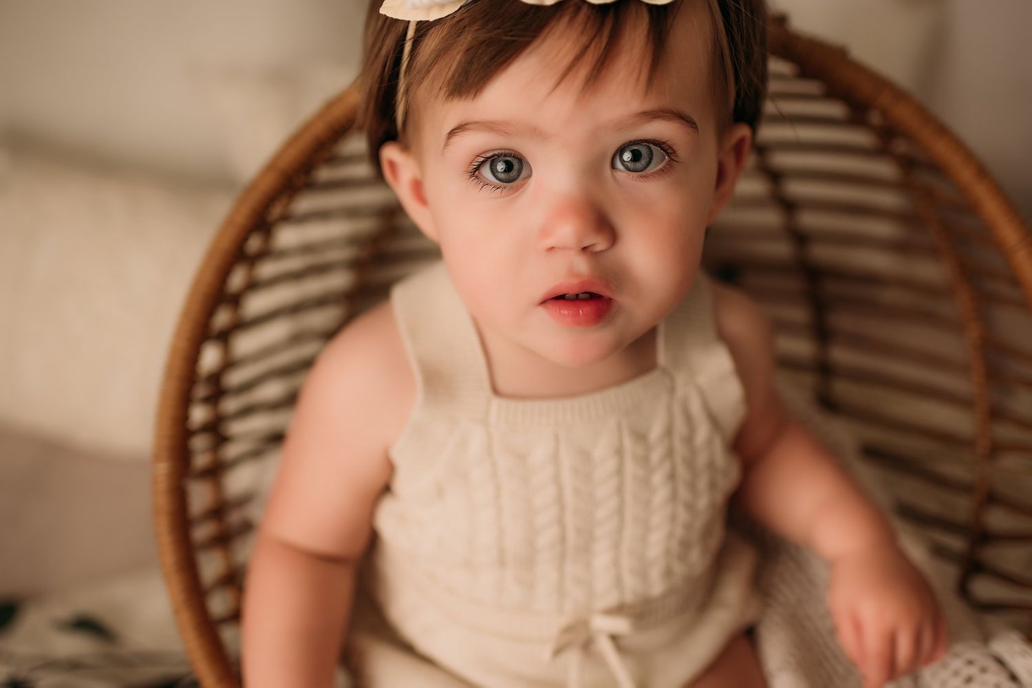 Close up portrait in rattan chair wearing cream knit romper with floral headpiece.