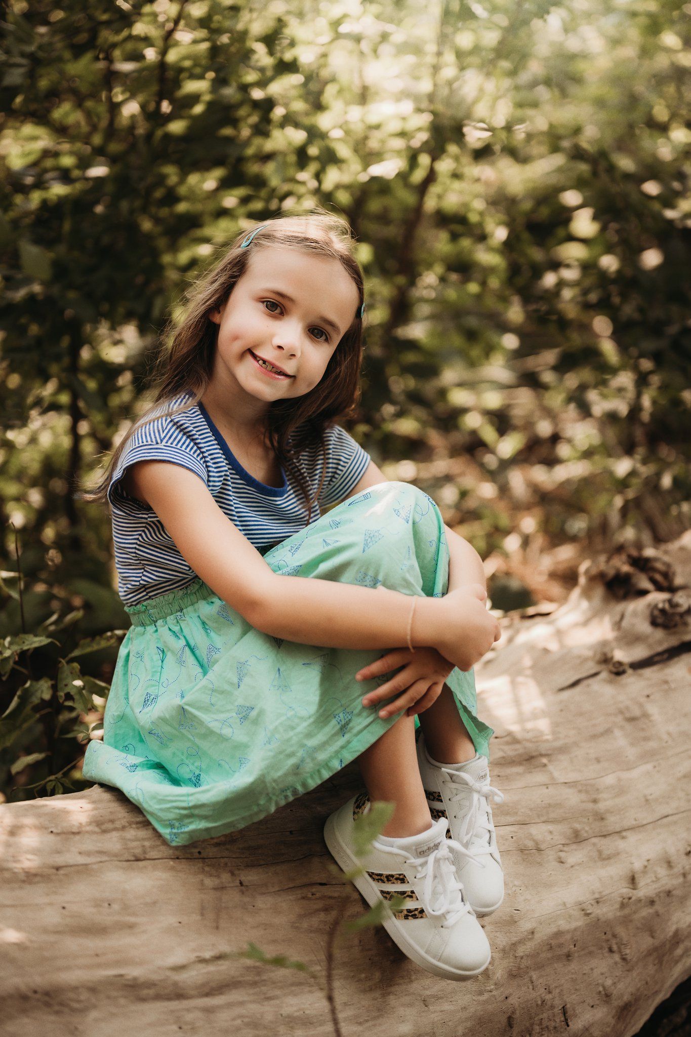 A young person in a striped shirt and mint green skirt sits on a log in a forest setting.