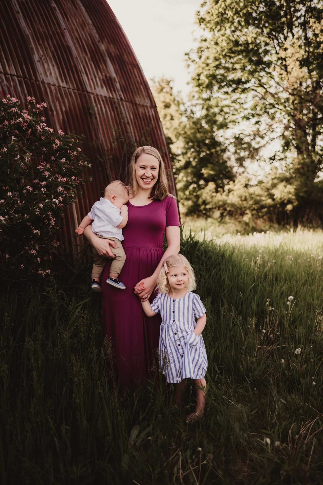 Family portrait by rustic barn with flowering bushes and natural summer lighting.