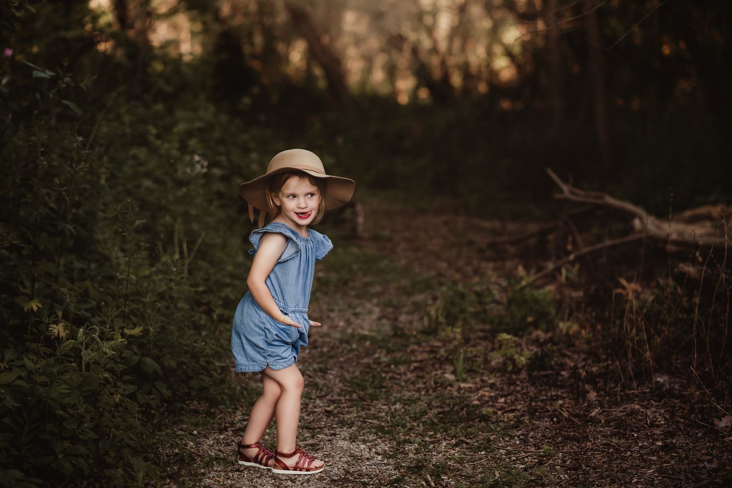 A playful pose in a blue romper and sun hat on a woodland trail.