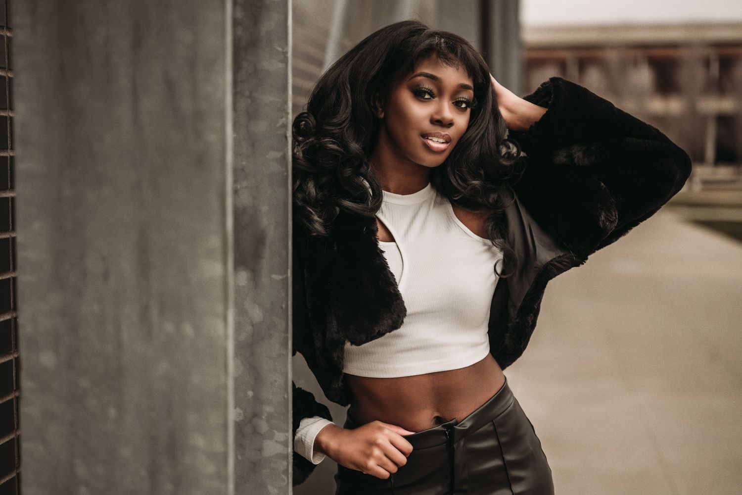 A fashionable model poses in a white crop top and black jacket against an urban backdrop.