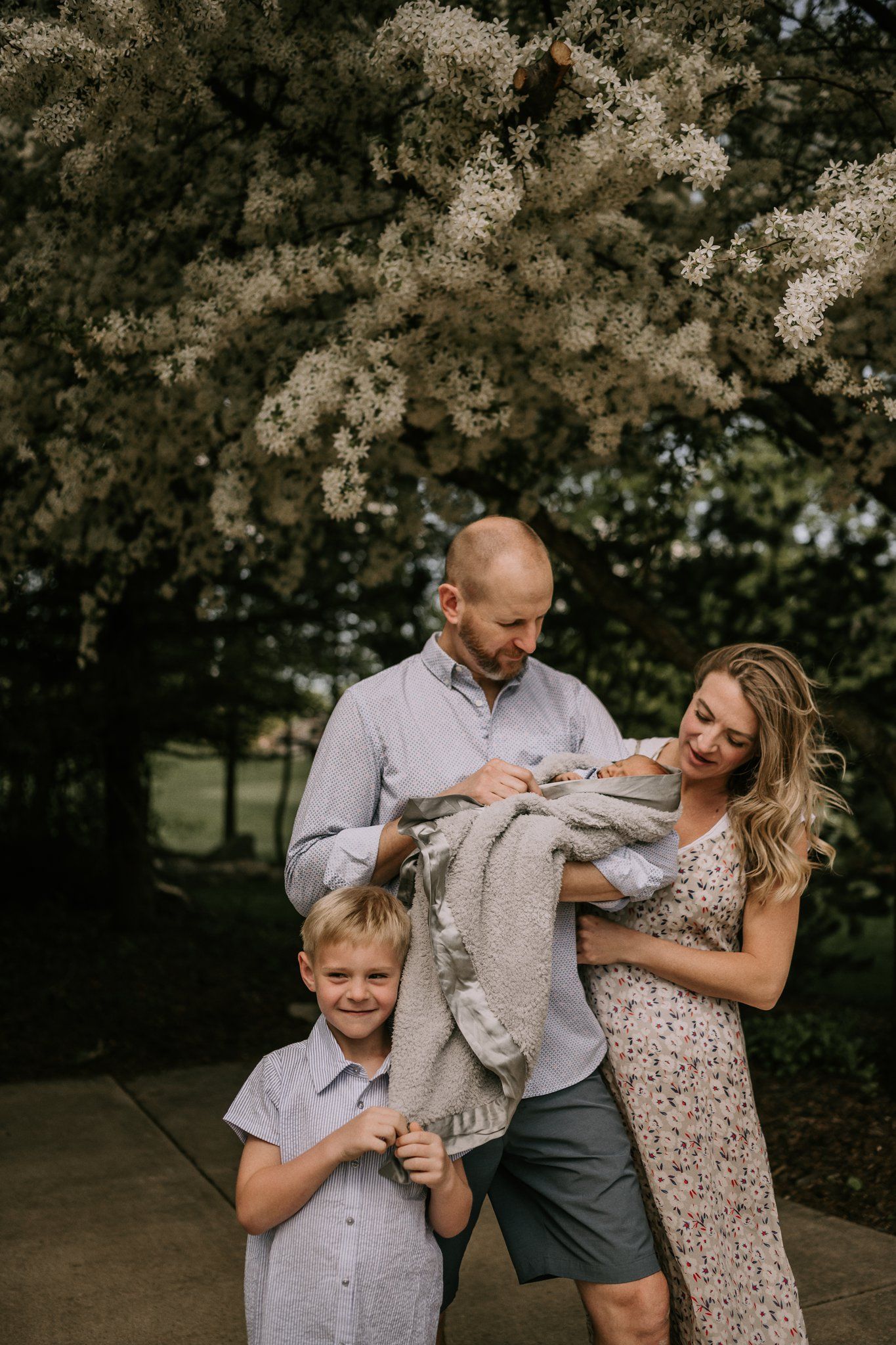 A family of three stands together under flowering trees while holding a swaddled newborn.