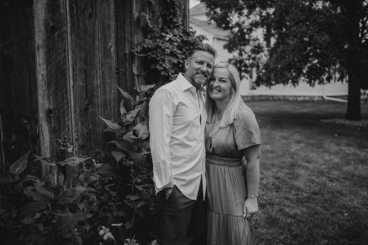 Black and white portrait of a couple laughing together against wooden barn background.