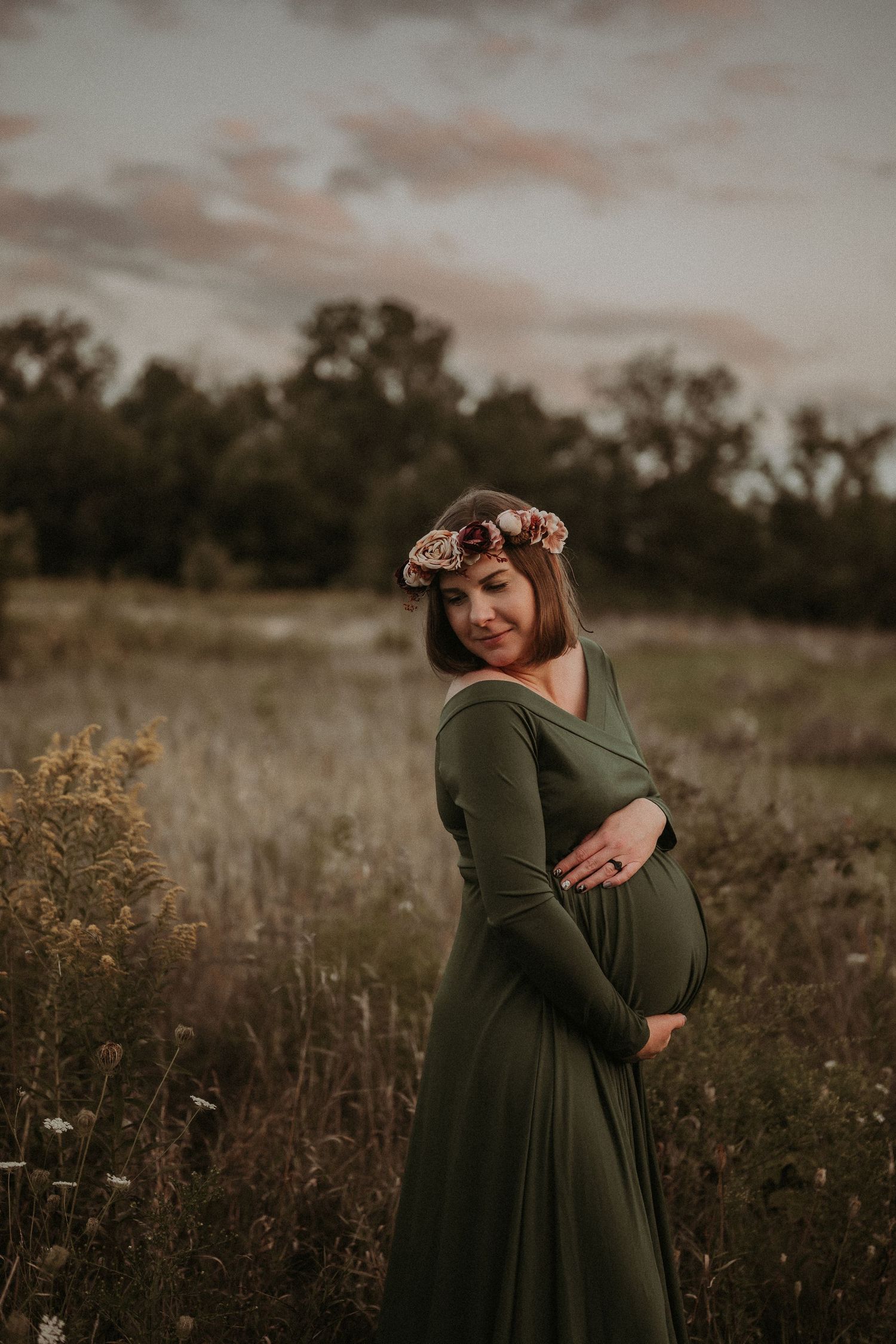 Maternity portrait in an olive dress with floral crown against moody sky.