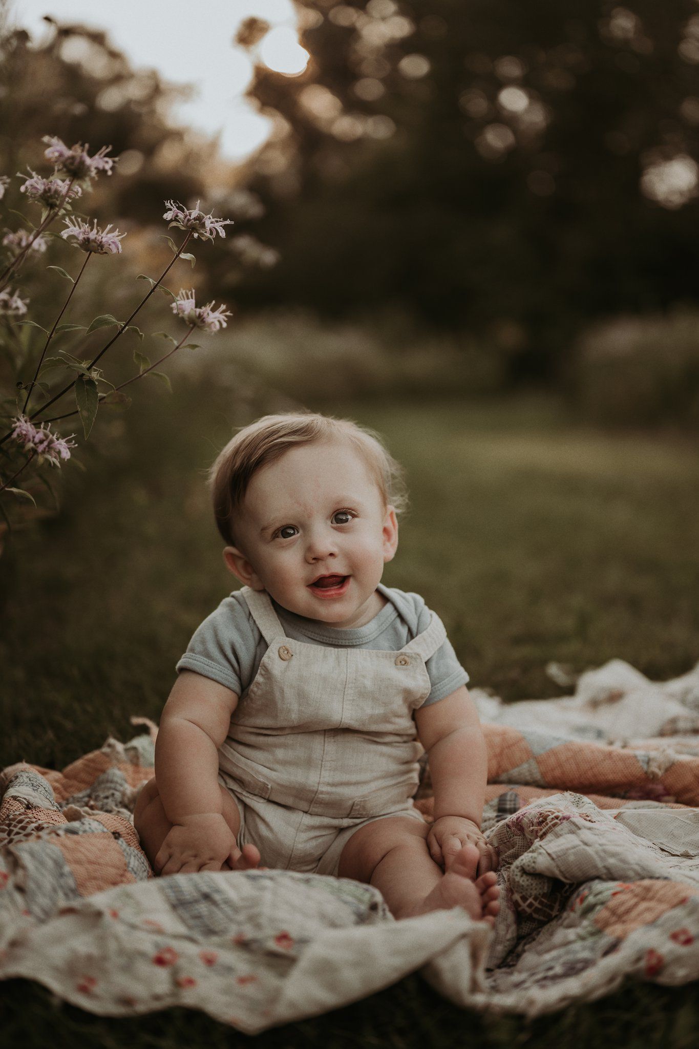 Baby in light colored overalls sits on floral blanket during golden hour outdoor photoshoot.
