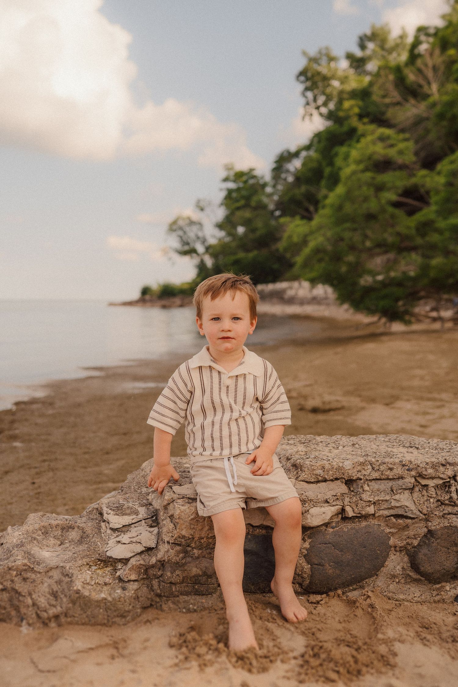 A small child sits on a rocky outcrop along a sandy beach during golden hour.