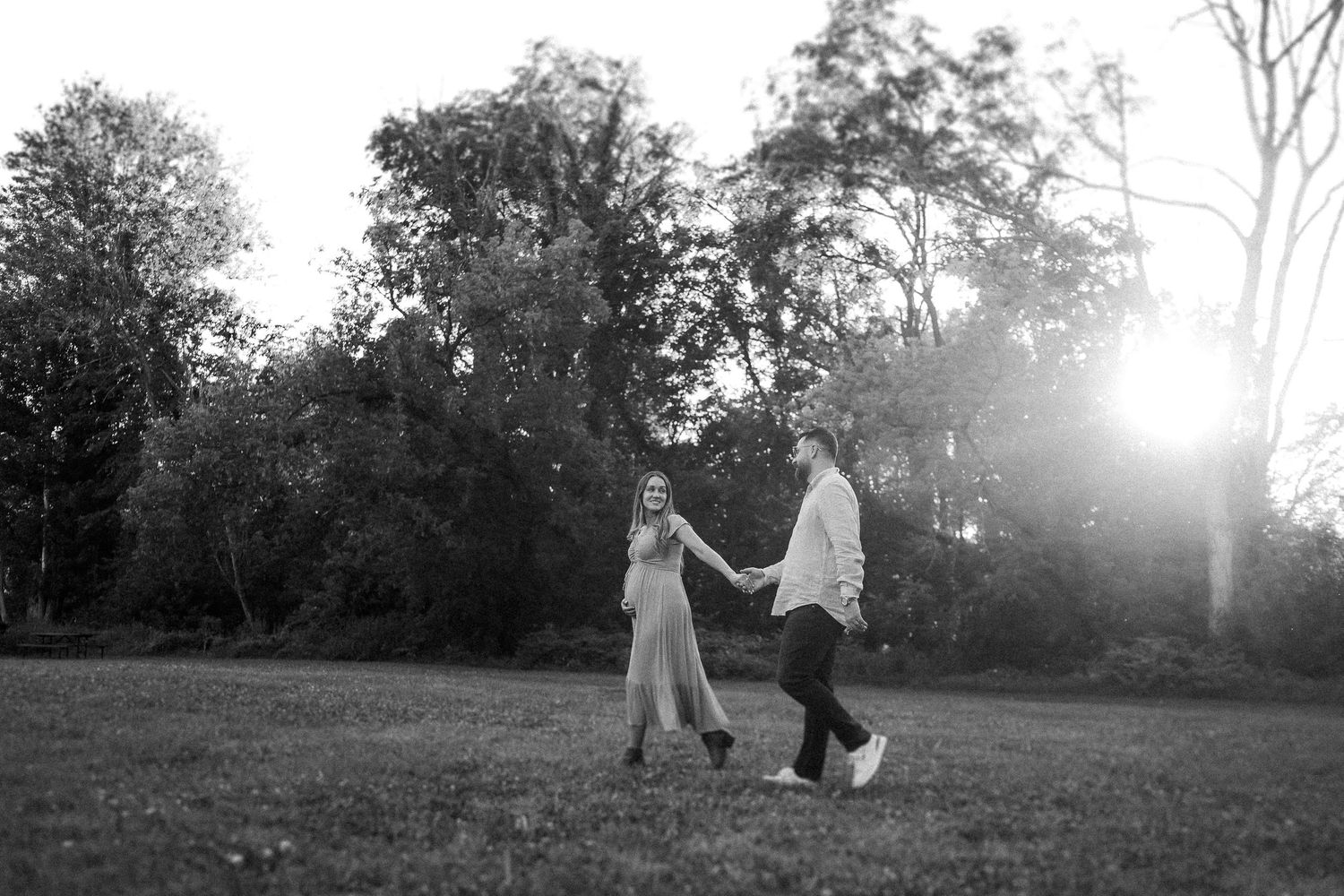 Two people dancing together in a grassy field at sunset with trees in the background, captured in black and white.