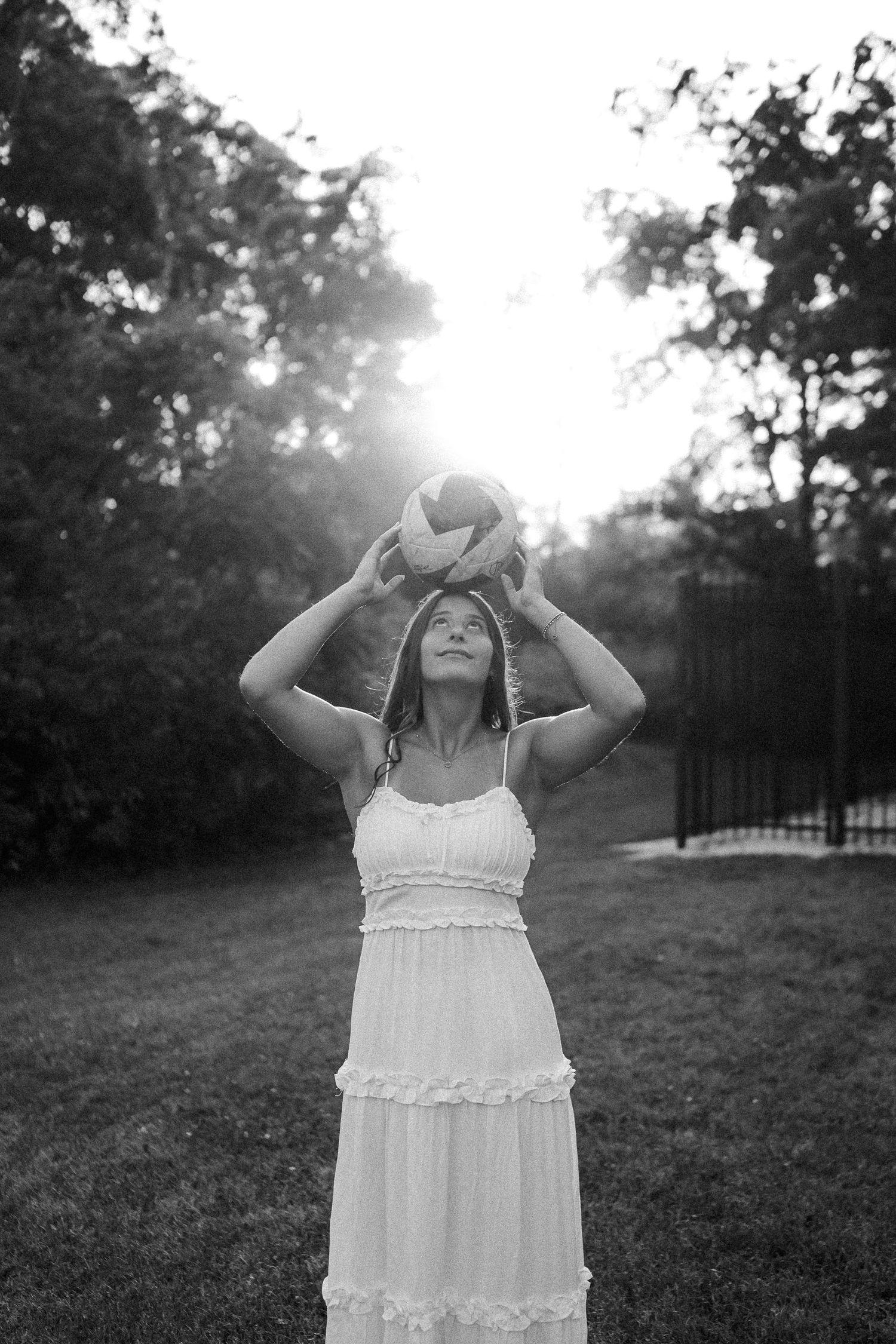 A person in a white tiered dress holds a volleyball overhead in a backlit outdoor setting.