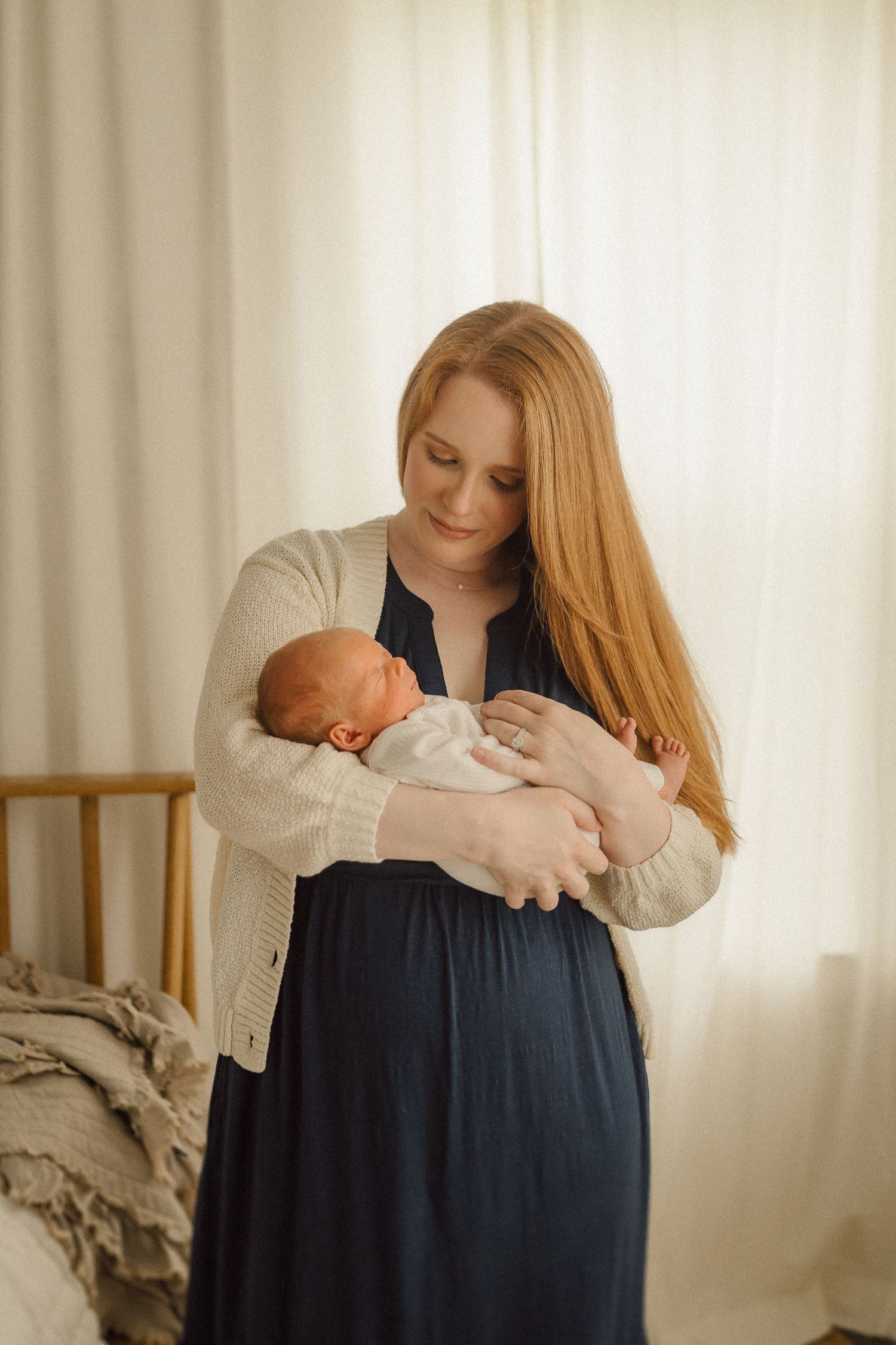 Mother in navy dress and beige cardigan tenderly holds sleeping newborn by bedroom window.