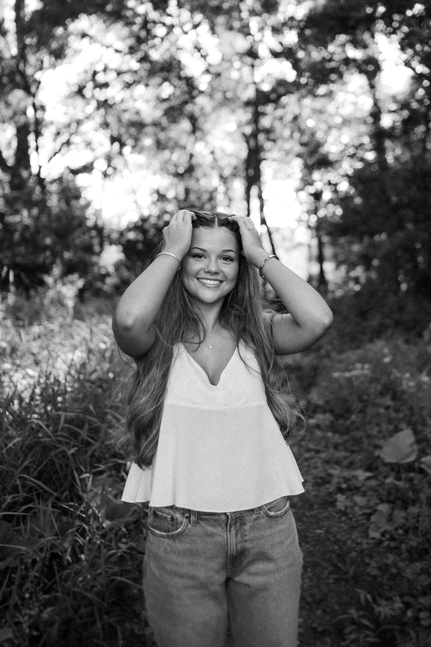 A joyful outdoor portrait in a forest setting featuring a white camisole top and denim jeans.