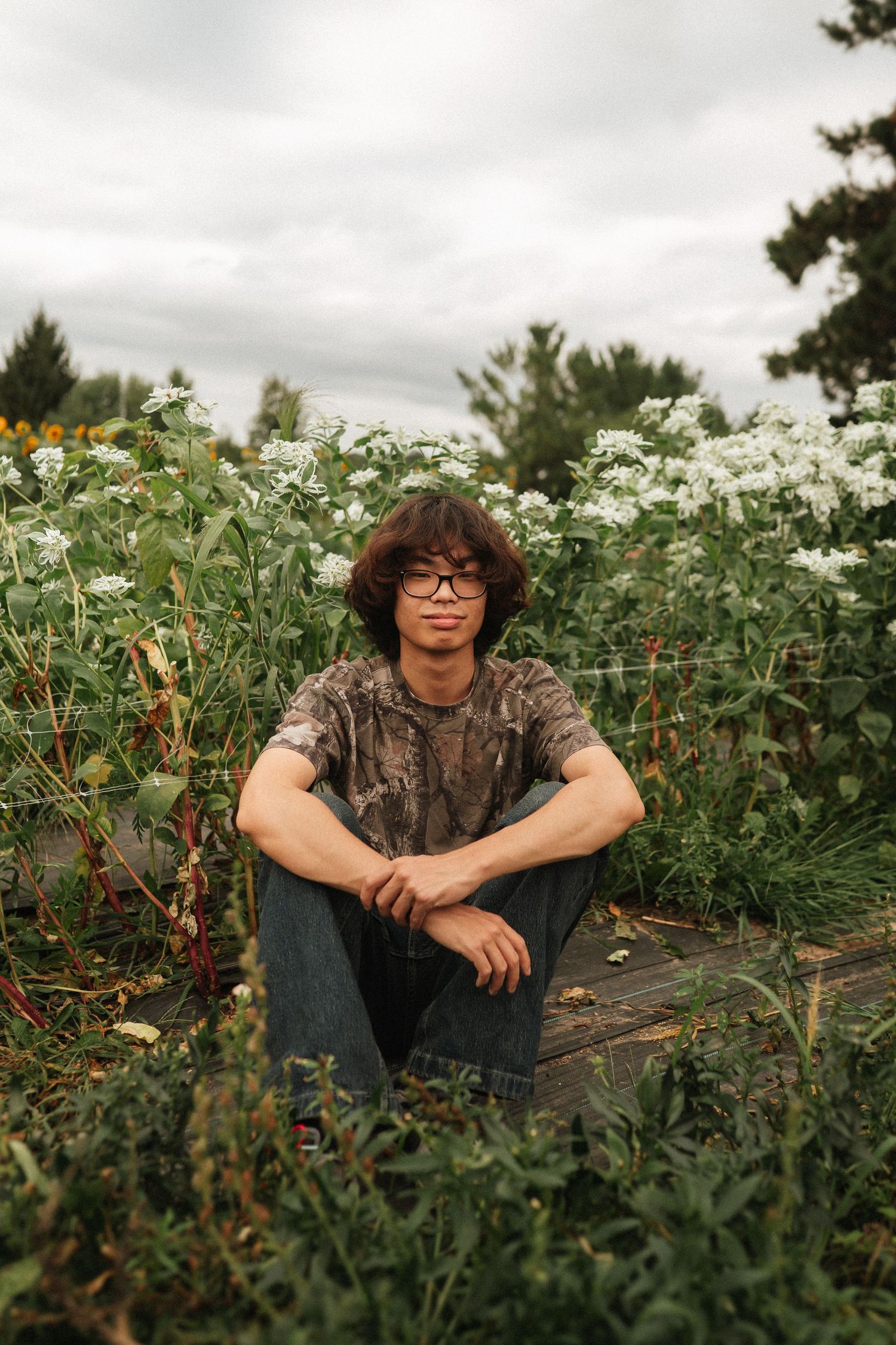 Someone wearing glasses and camouflage sits among green plants and white wildflowers on an overcast day.