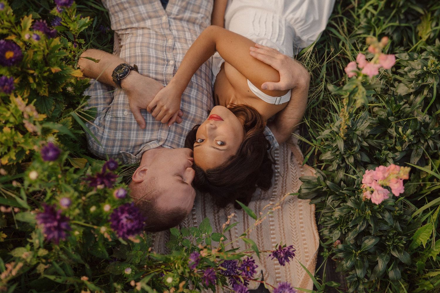 Person lying on a picnic blanket surrounded by purple flowers and greenery in a romantic garden setting.