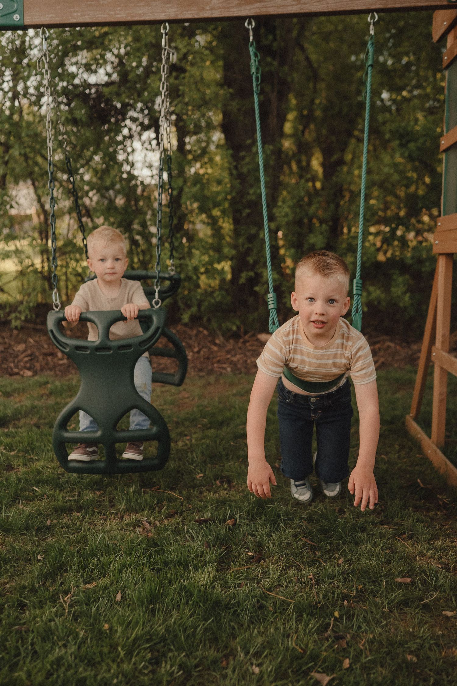 Two young boys play on green chain swings in a backyard playground surrounded by lush green trees.