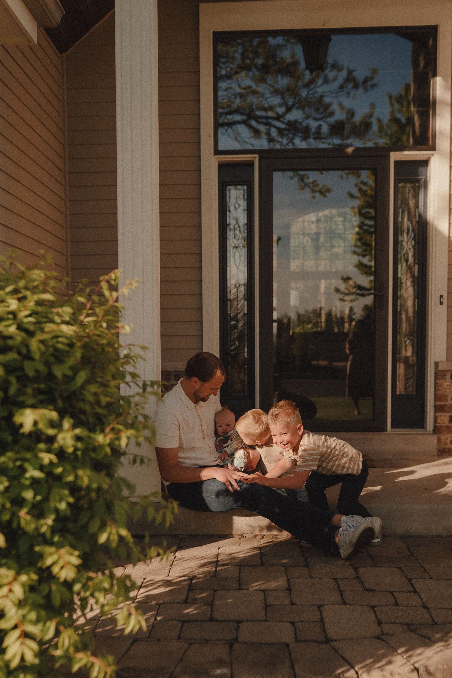 A father sits on a sunny front porch with two young children, sharing a moment together outside their home.