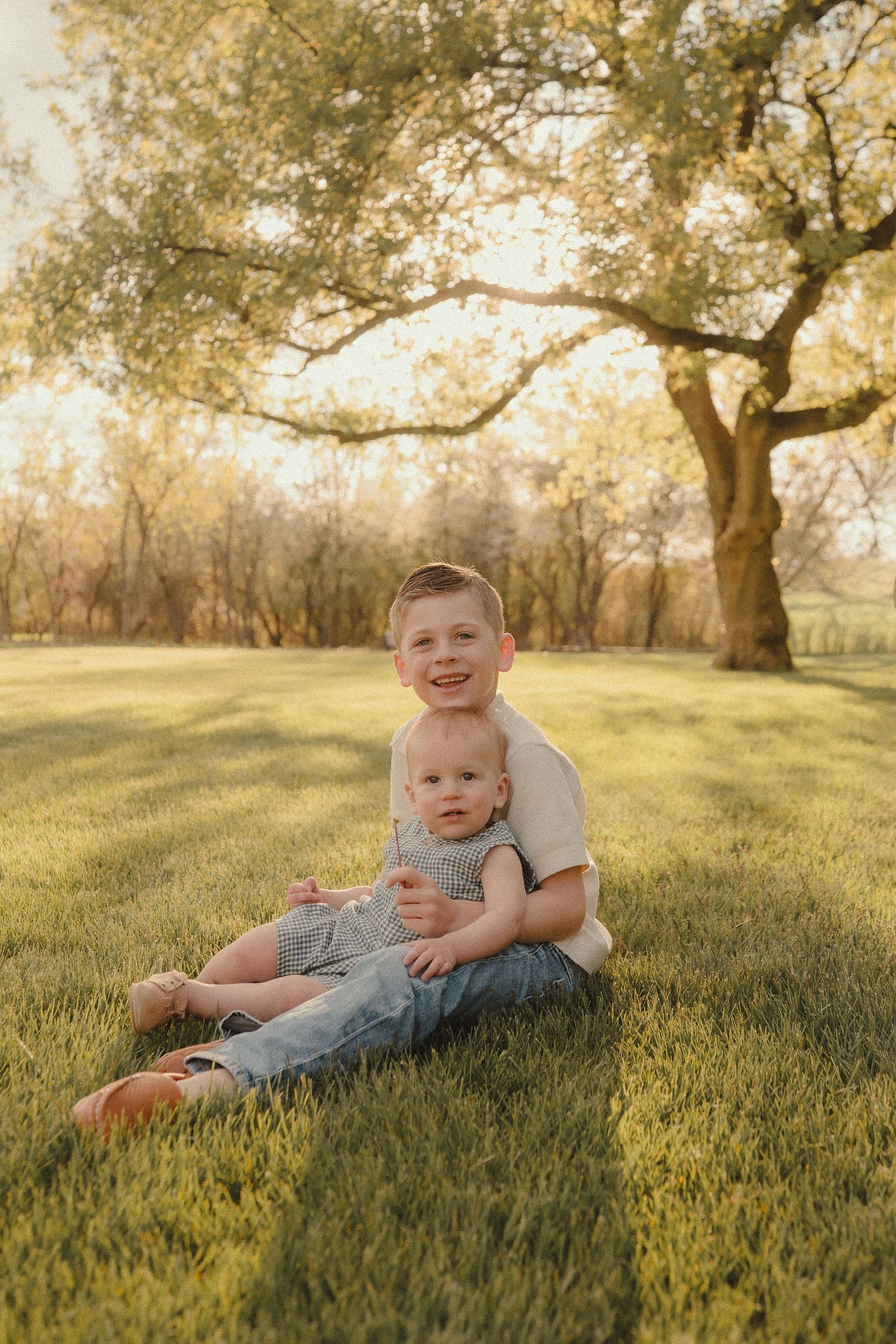 A smiling boy sits on green grass holding a baby sibling under a golden sunlit tree outdoors.