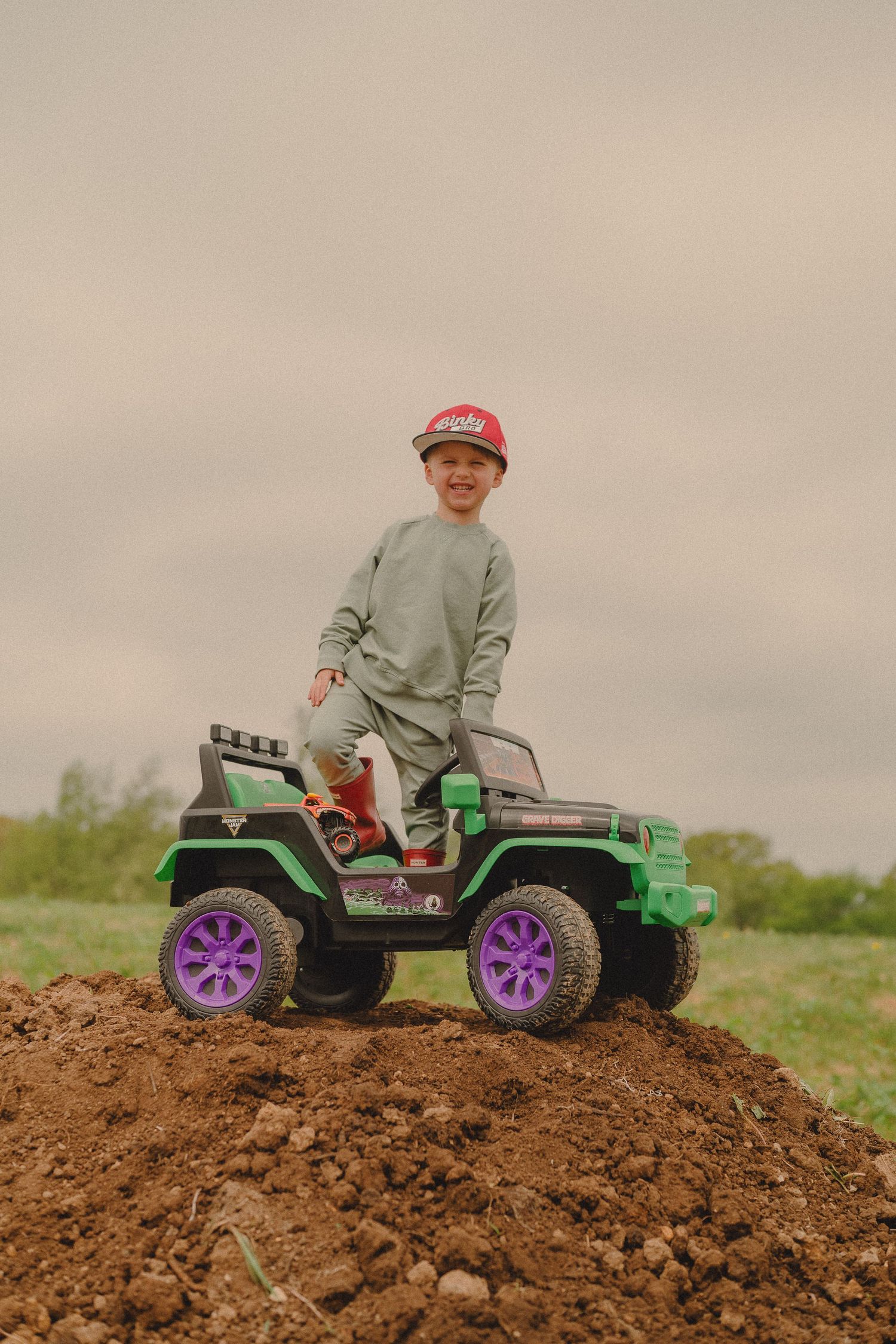 Happy boy in red cap sits atop green ride-on monster truck jeep on dirt mound.