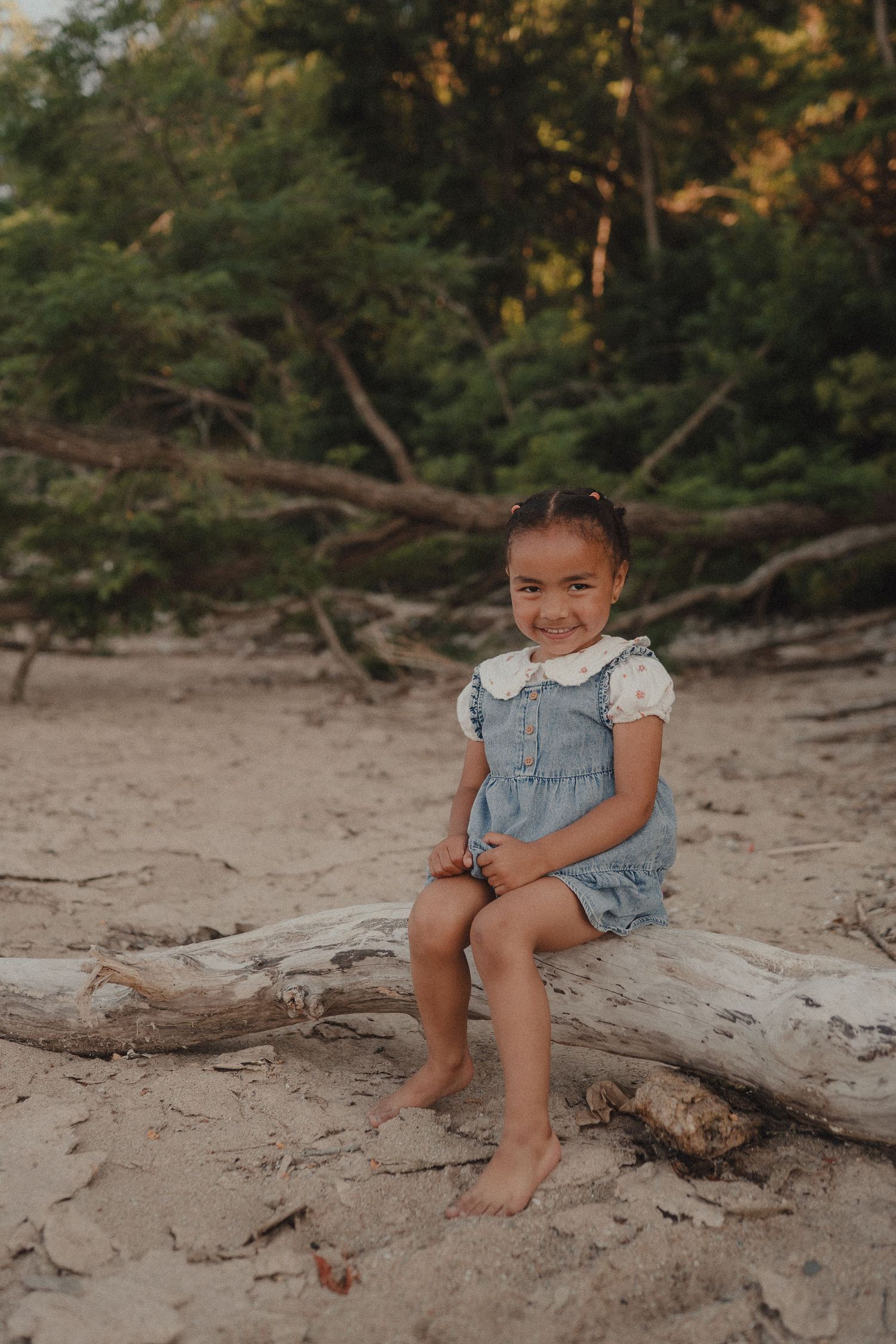 A smiling young girl in a blue denim romper sits barefoot on driftwood at a sandy beach with green trees behind her.