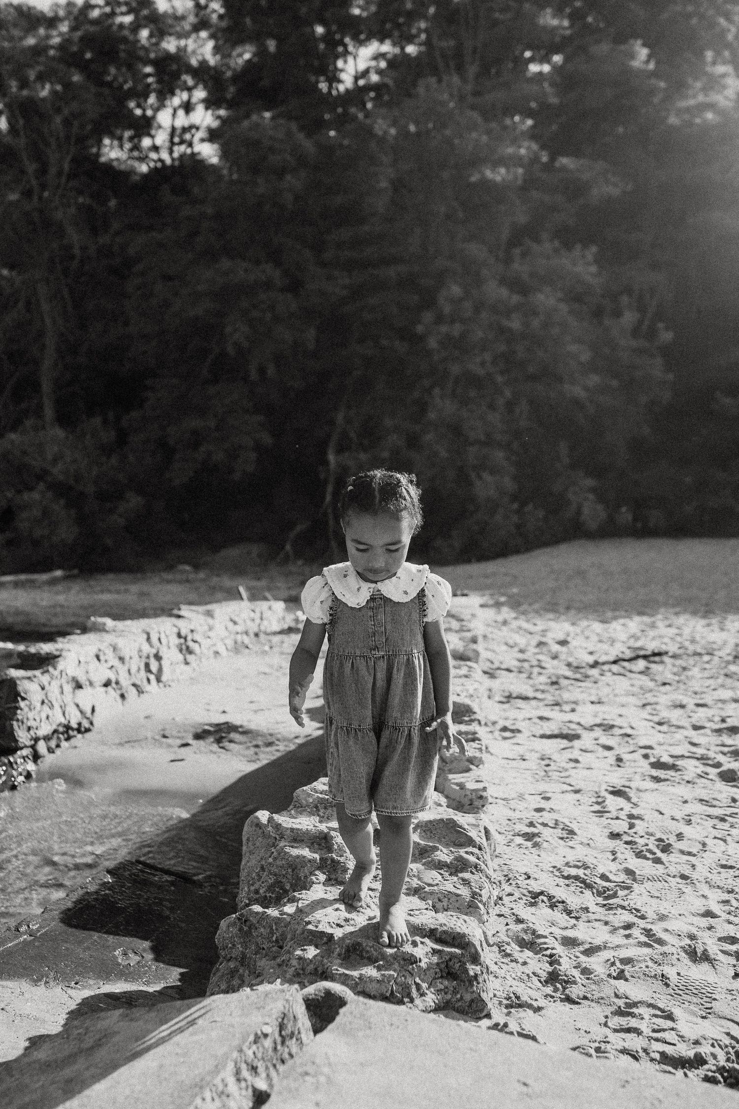 A black and white photo of a toddler girl in a ruffled dress walking on rocks near a sandy beach.