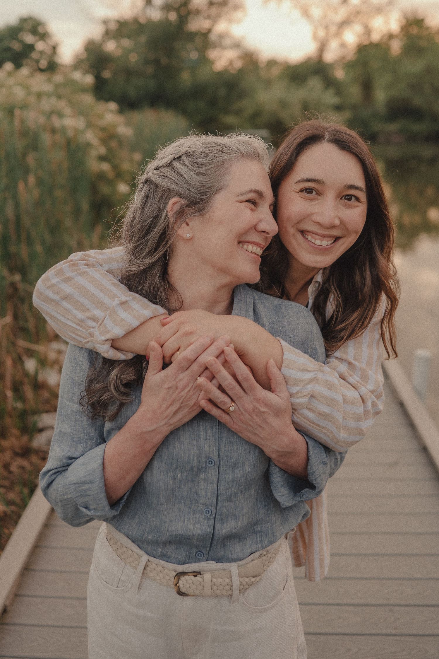 A younger woman hugs an older woman from behind on a wooden dock, both smiling warmly outdoors.
